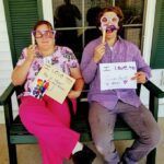 Two people holding props, wearing glasses and holding signs on a porch. One in pink, the other in purple, expressing love.