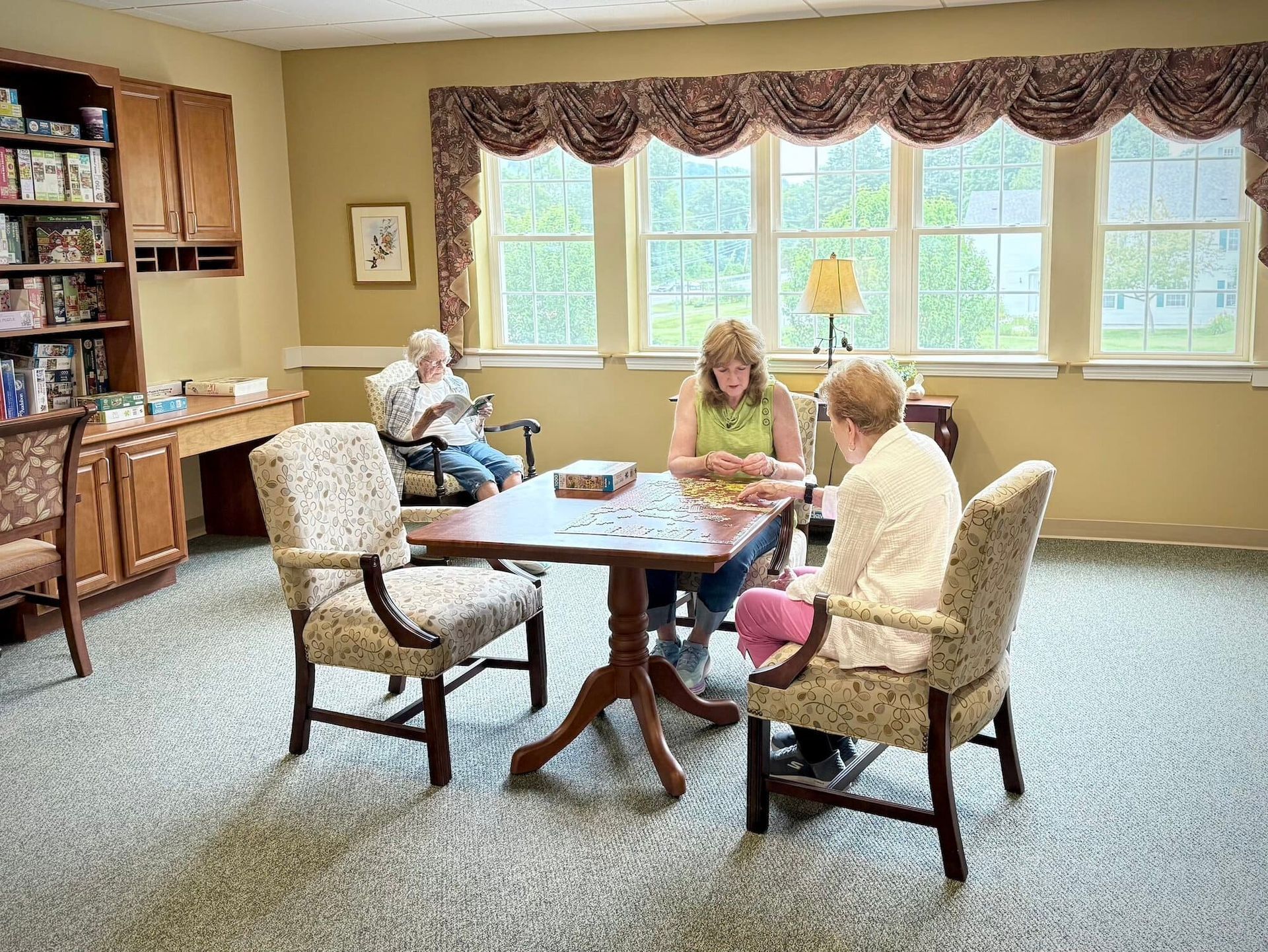 Three people seated around a table, one in a wheelchair, doing a puzzle in a room with a bookcase and large window.