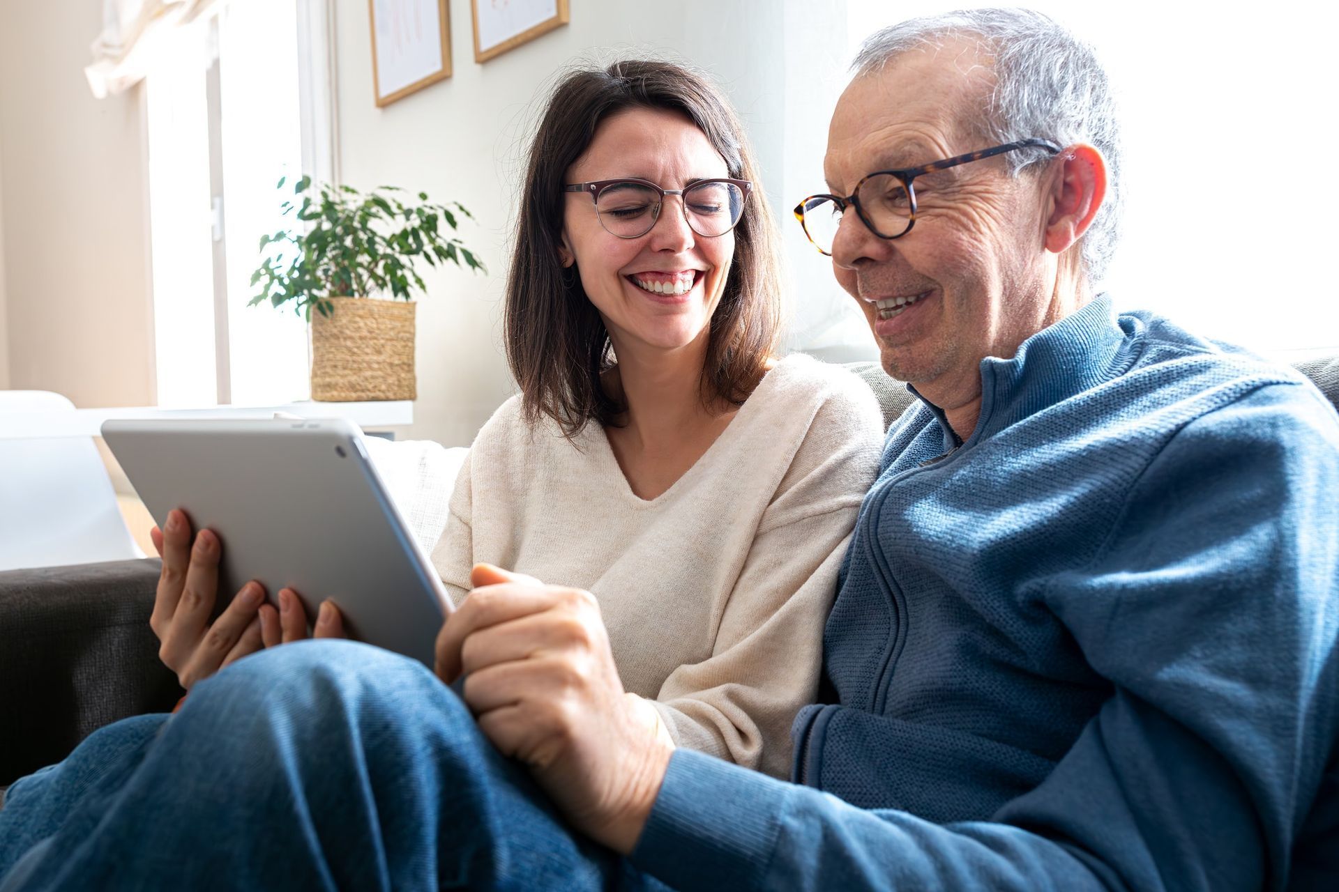 Woman and older man looking at tablet together, both smiling. Sunlight streams in.