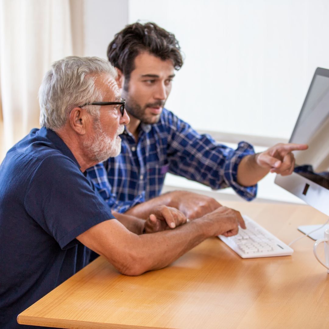 Man points at computer screen, assisting another man with keyboard; indoor, daytime.