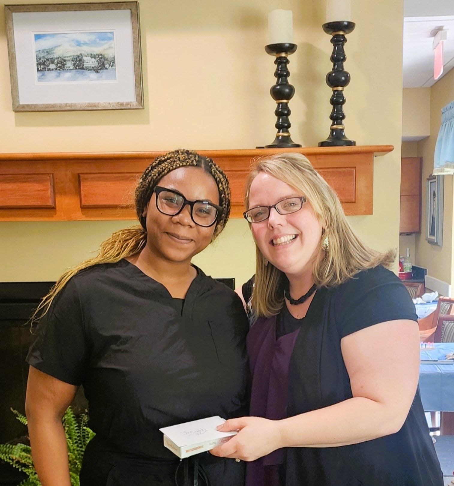 Two women smiling and holding a small gift box near a mantelpiece with candles and artwork.
