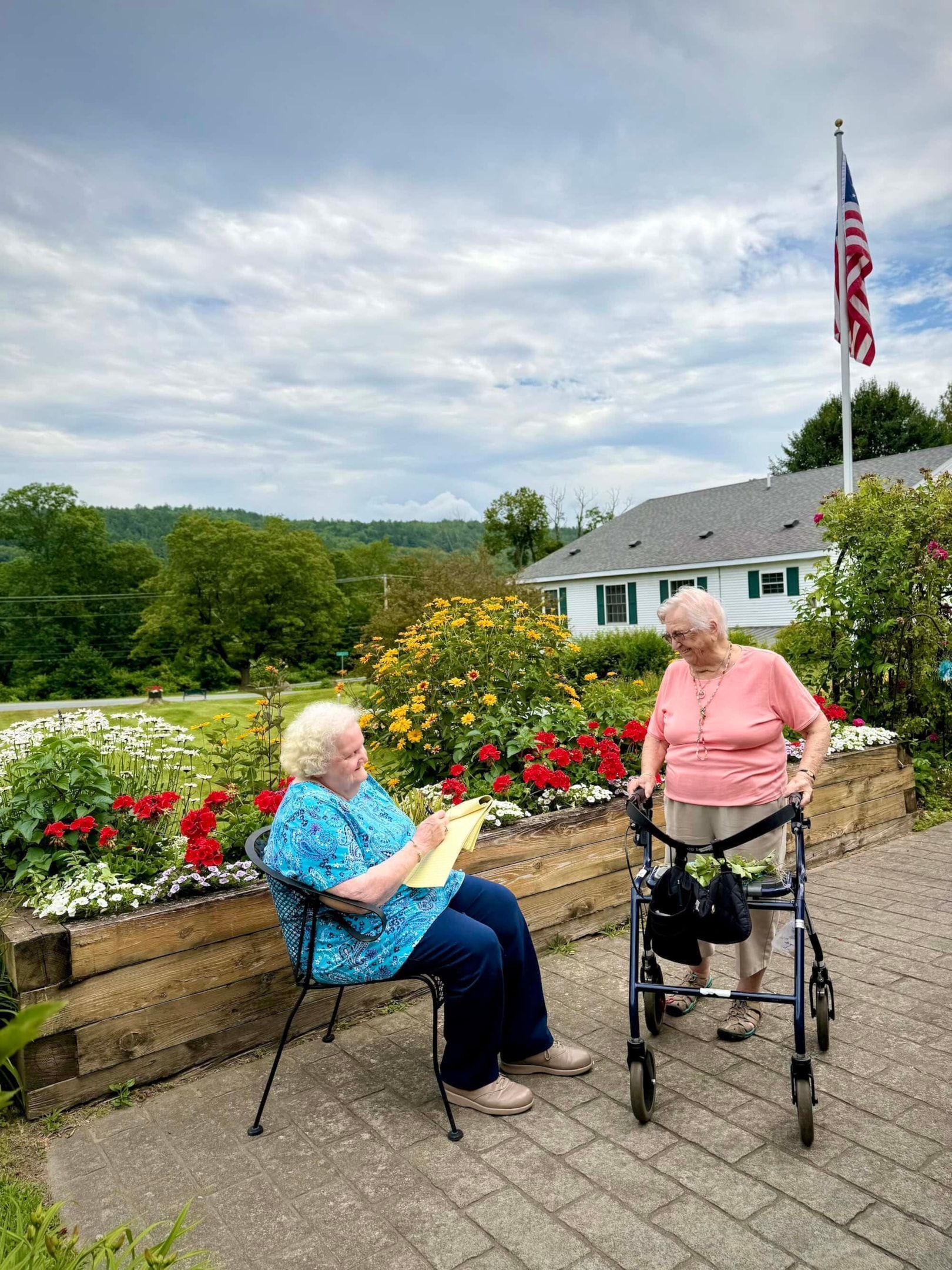 Two women outdoors near a flower bed and house. One sits, writing; the other uses a walker. An American flag flies.