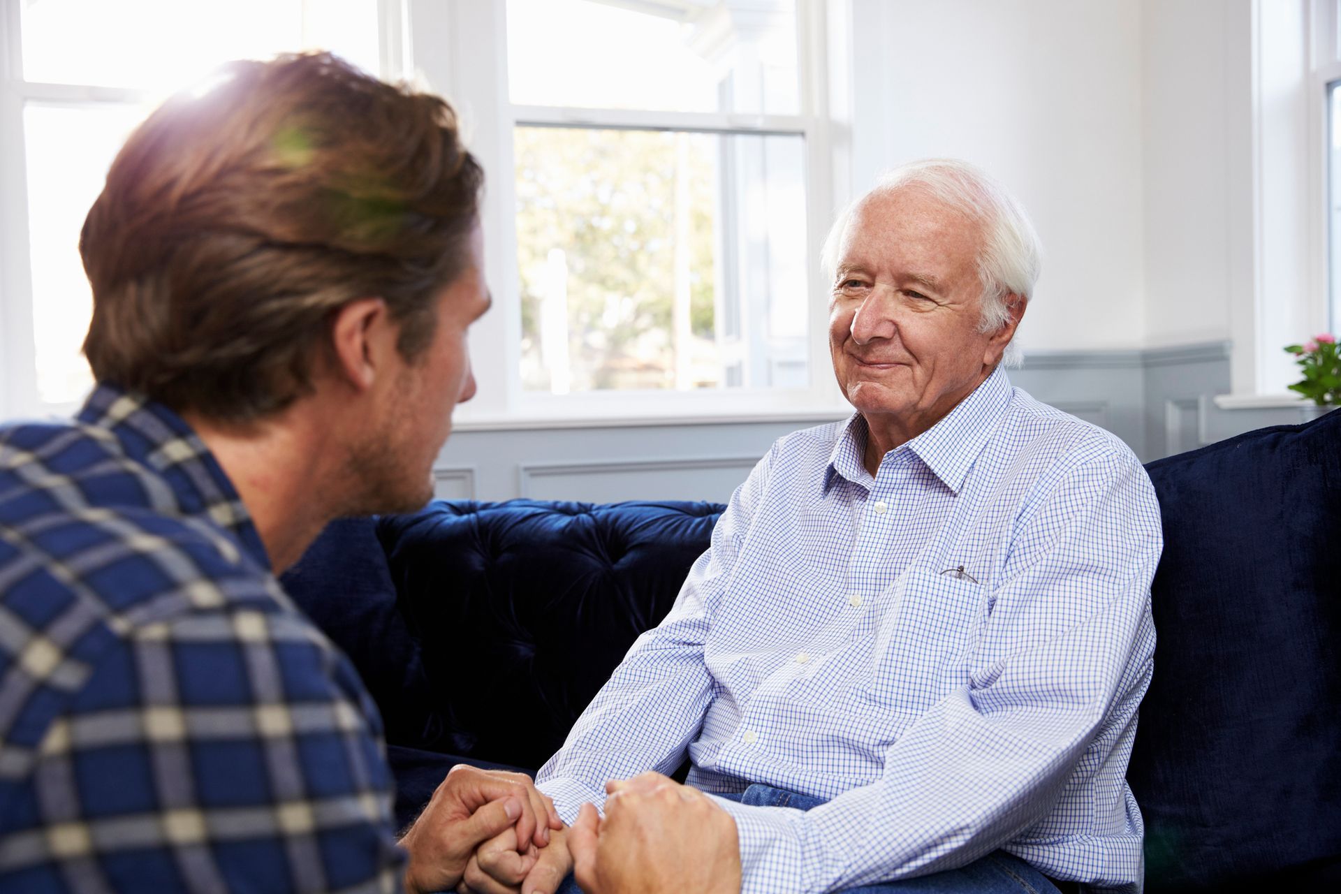 Man holding hands with an older person on a couch, indoors, both smiling.
