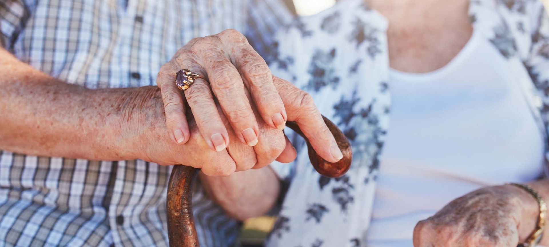 Elderly couple holding hands, one with ring, resting on a cane, wearing patterned shirts.