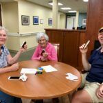Three smiling seniors at a round table, holding phones up. A light-filled room with artwork on the wall.