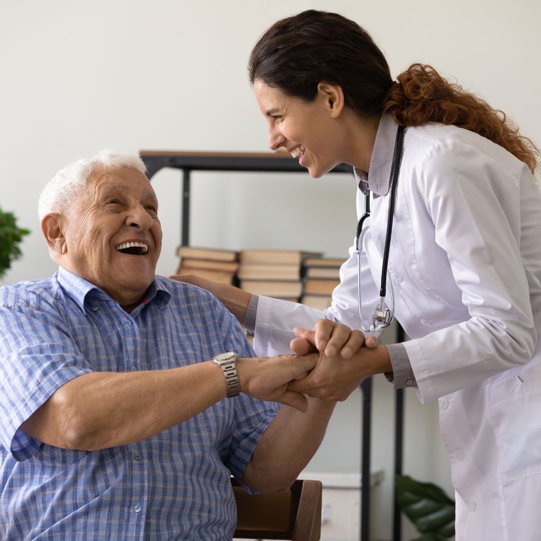 Elderly man smiles, holding hands with a smiling doctor in a white coat. Indoor setting.