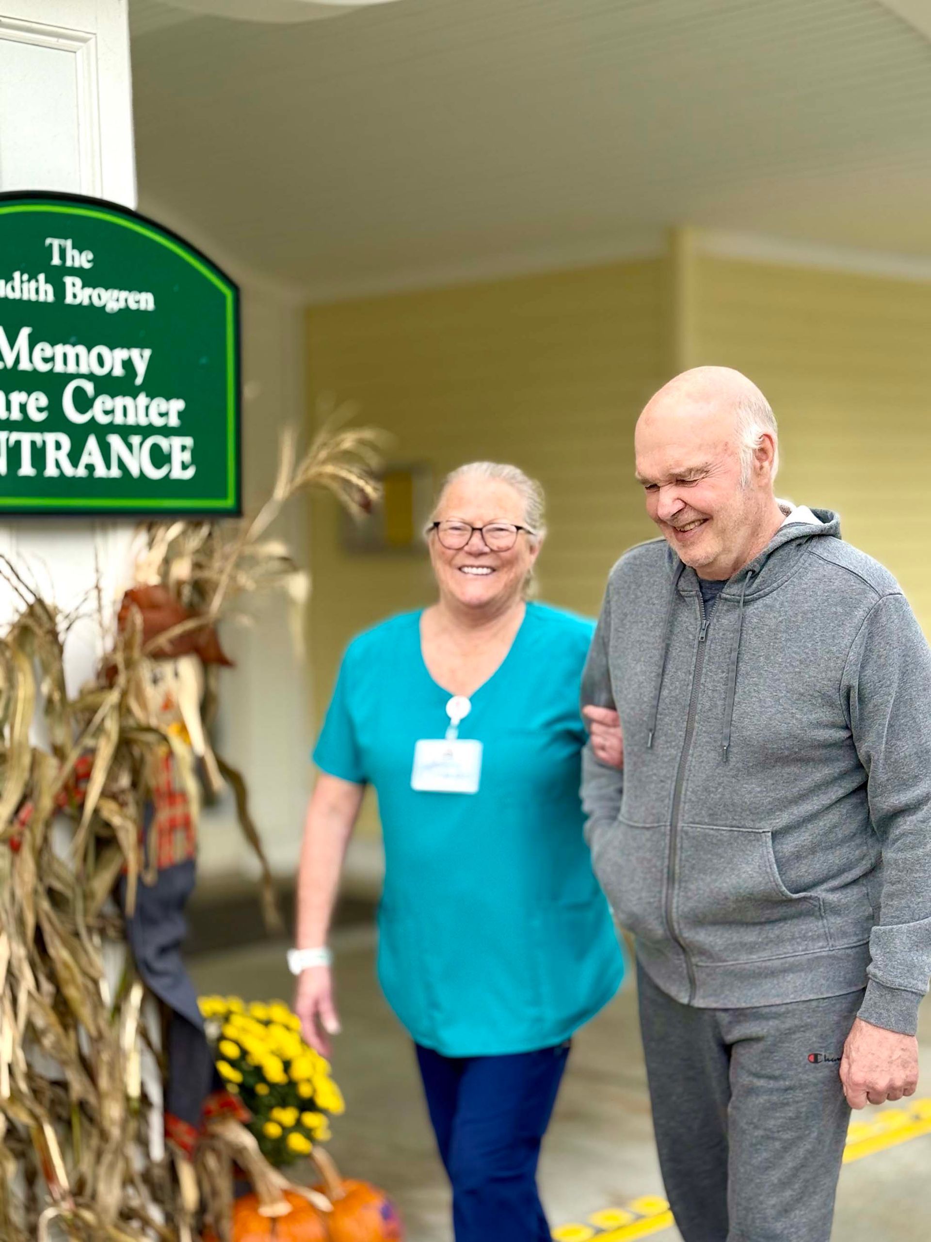 A care worker in a teal uniform walks with a smiling resident outside the entrance of a memory care facility.