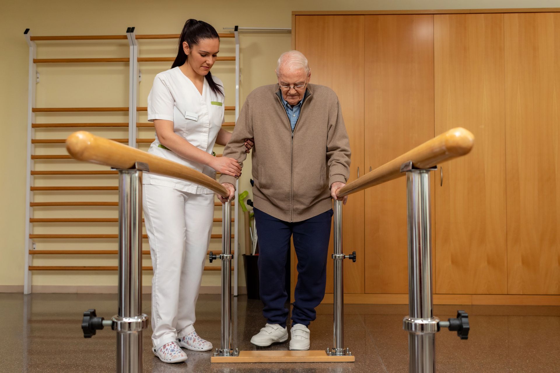A healthcare worker assists a person walking between parallel bars in a therapy room.