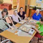 Group of seniors and caregivers baking cookies at a table; indoors, smiling faces, wearing gloves.