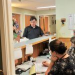 Man speaking at front desk, two women attend. Office setting with light colors, paperwork visible.