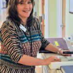 Woman with brown hair smiles, wearing patterned dress, leans on a counter.