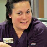 Woman in purple scrubs smiles, name tag visible.