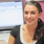 Woman smiles at camera; dark hair, black shirt, computer keyboard in the background.