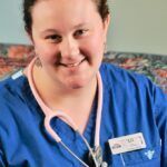 Woman in blue scrubs with pink stethoscope, smiling, in a medical setting.