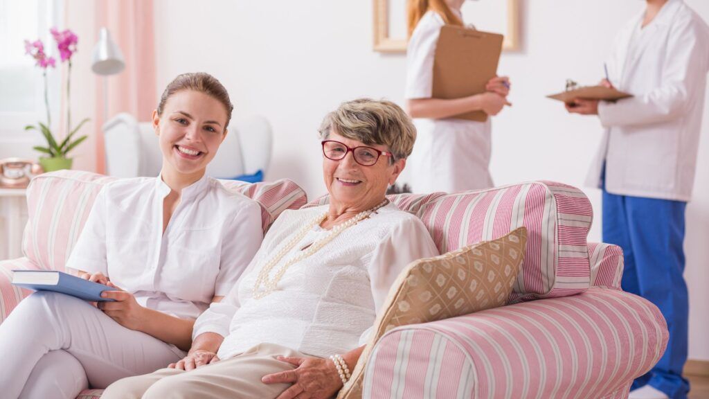 Woman and caregiver smiling on a couch, two nurses in background. Pink striped couch in room.