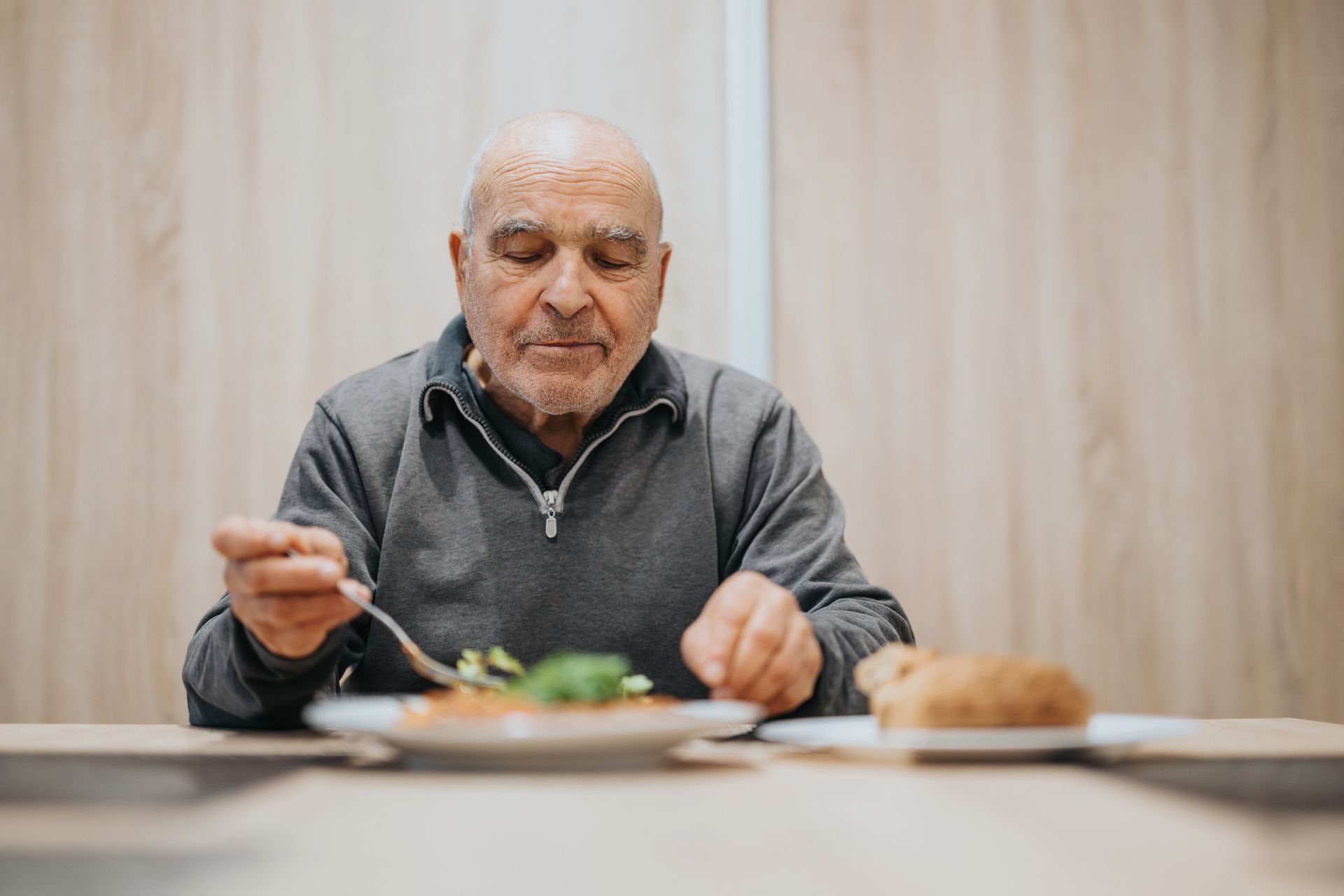 Older person eating a meal at a table, focused expression, plate of food and bread.