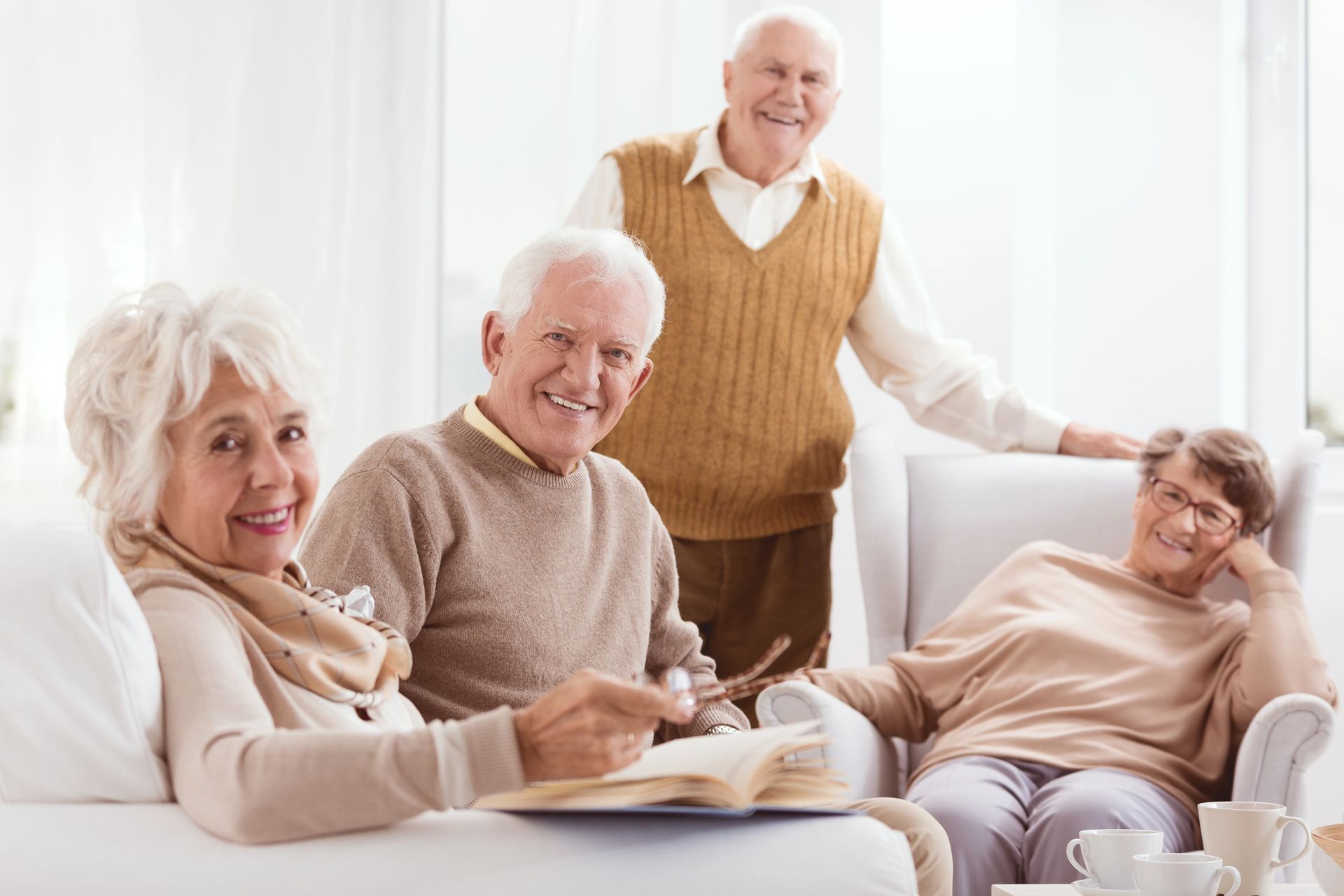 Four older adults smiling together, relaxing indoors; two seated, two standing.