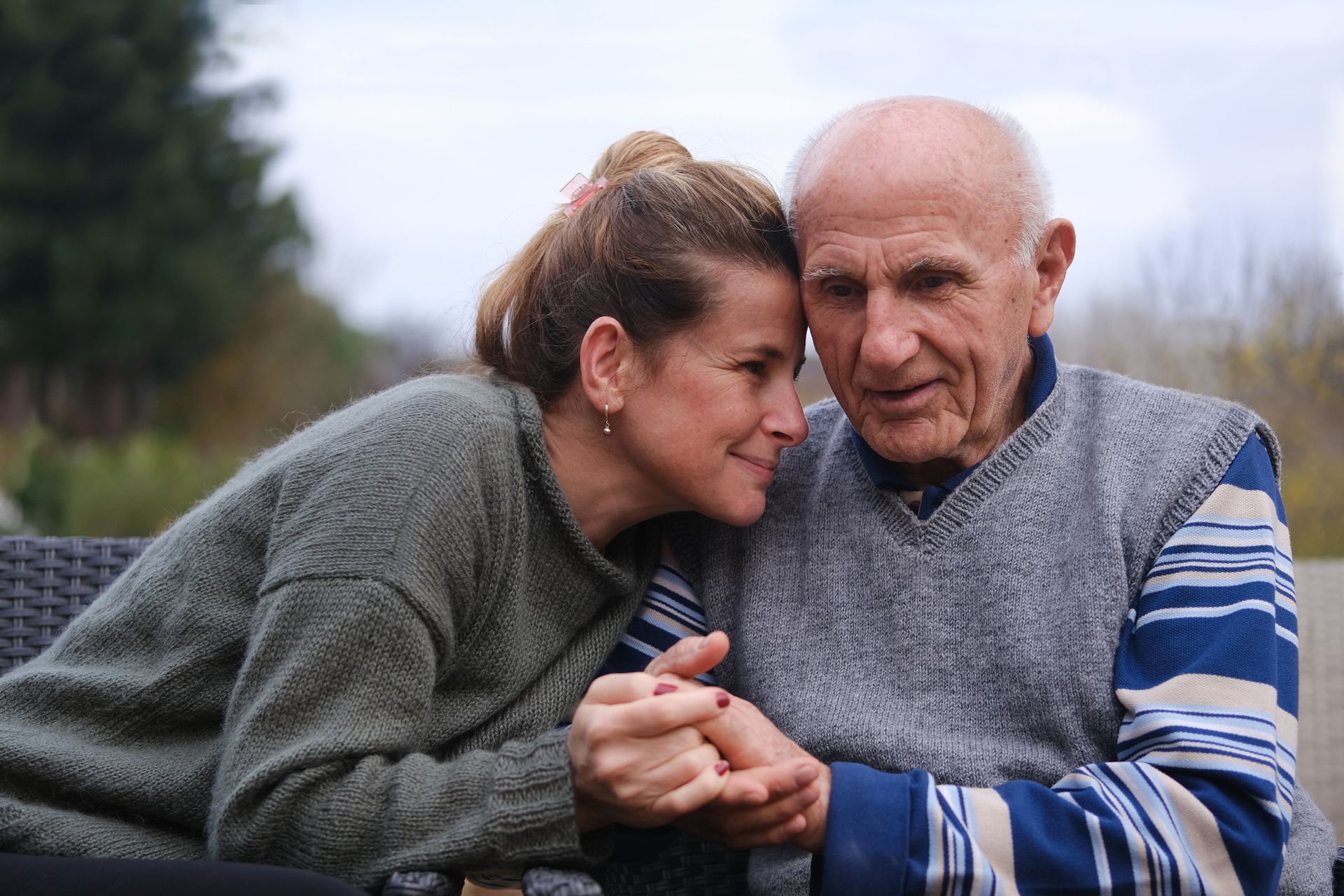 Woman and elderly man, holding hands, looking at each other with care. Outdoors, overcast sky.