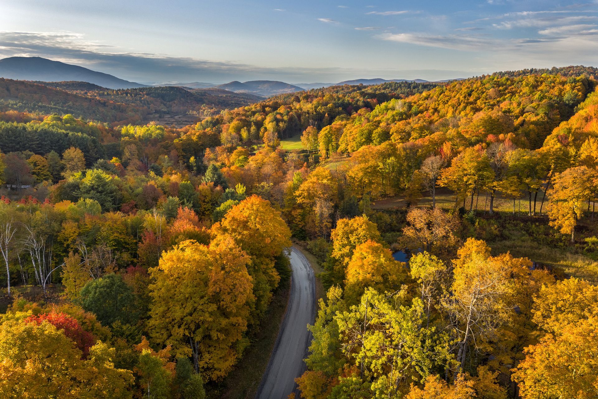 A winding road through a vibrant autumn forest, with golden, orange, and green foliage covering the hills.