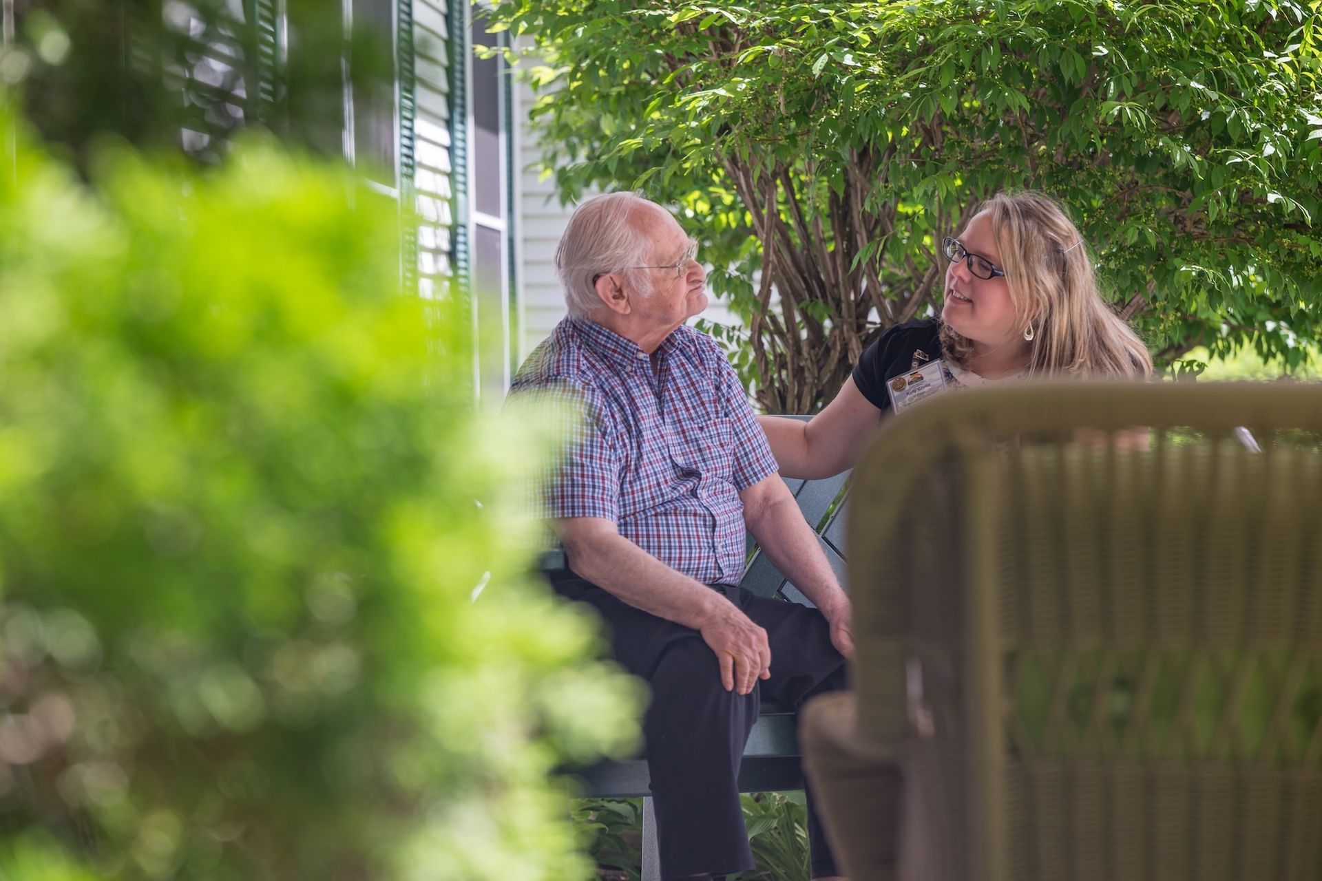 An older person and a younger person sit on a porch, interacting. The younger person has a hand on the other's shoulder.