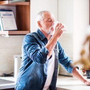 An older adult drinks from a glass in a kitchen, wearing a blue shirt.
