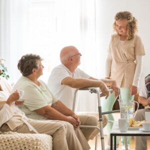 Caregiver with elderly people in a light-filled living area. They are socializing, with a walker nearby.