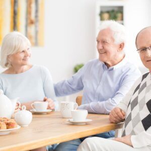 Three older adults at a table, smiling while having tea and snacks.