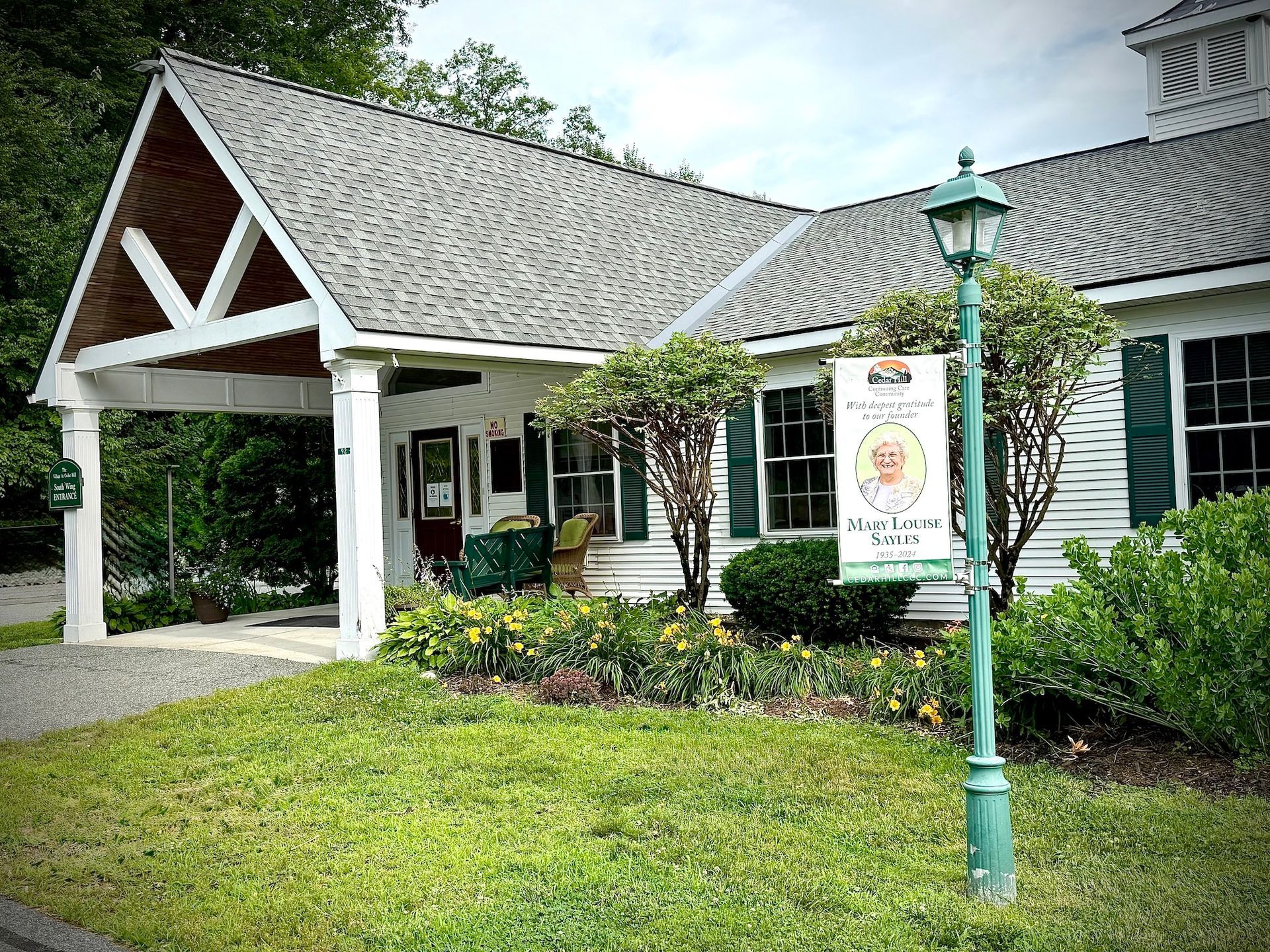 White cottage-style storefront with green trim, porch, sign, and landscaped lawn under a cloudy sky