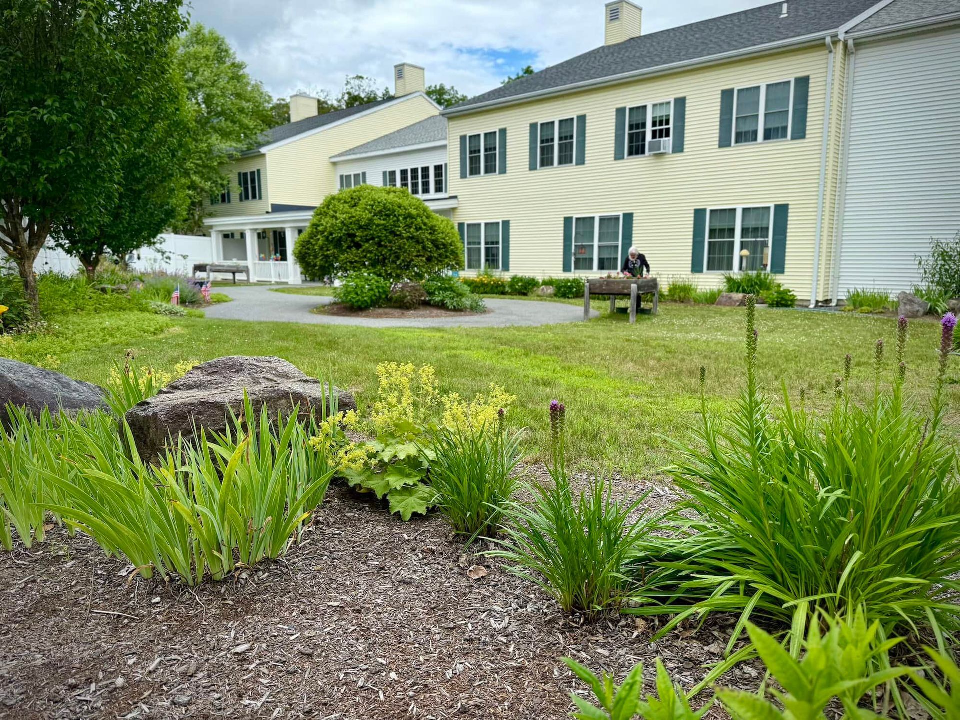 Lawn and flowerbeds in front of a light yellow building with green shutters; overcast sky.