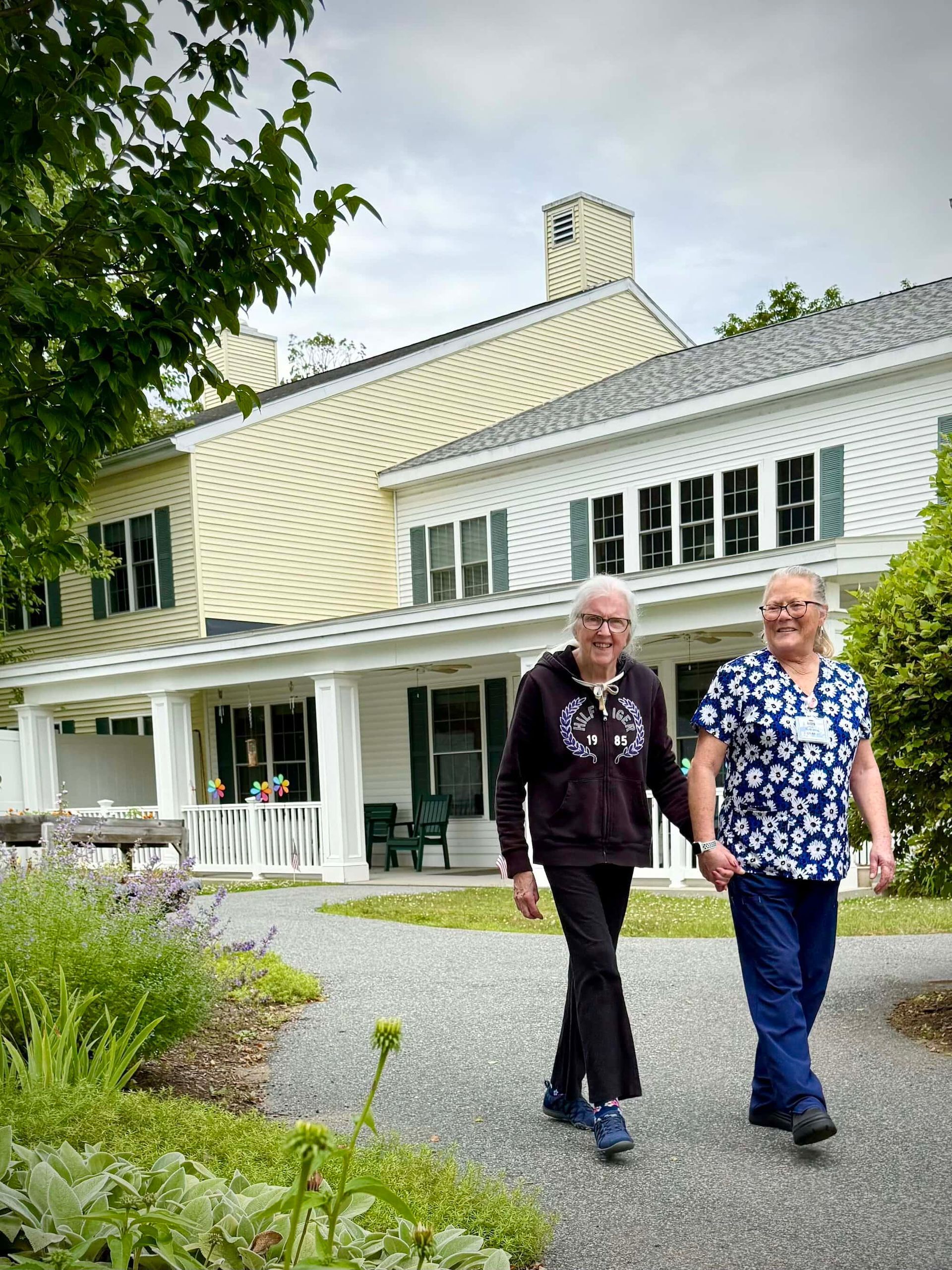 Two people holding hands walking on a path in front of a white house with a yellow roof.