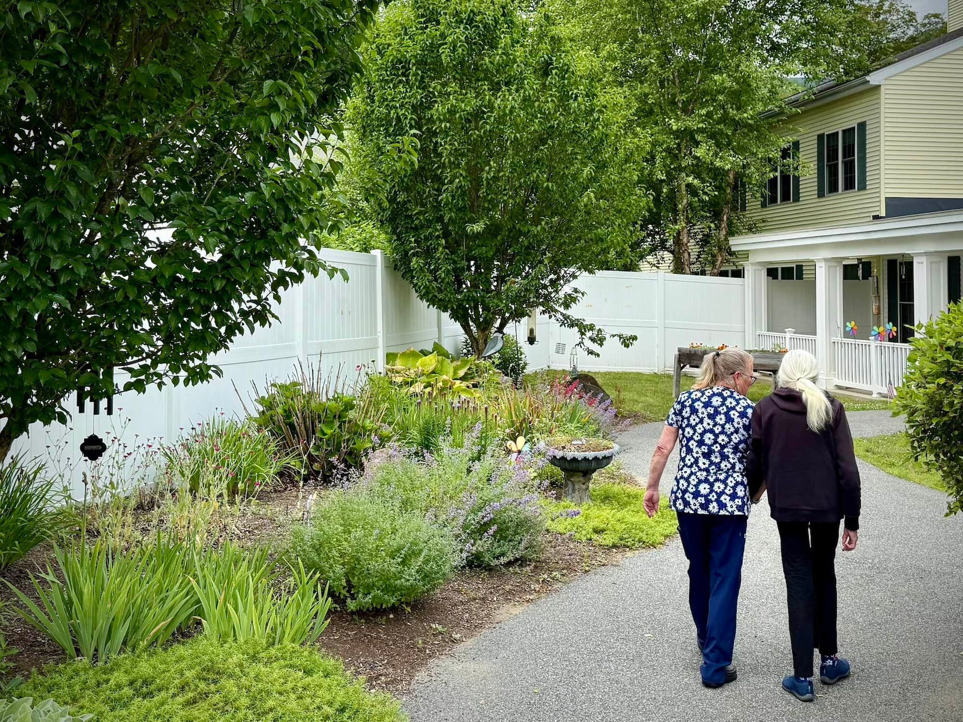 Two people walking on a gravel path in a garden, with a white fence and a building visible.