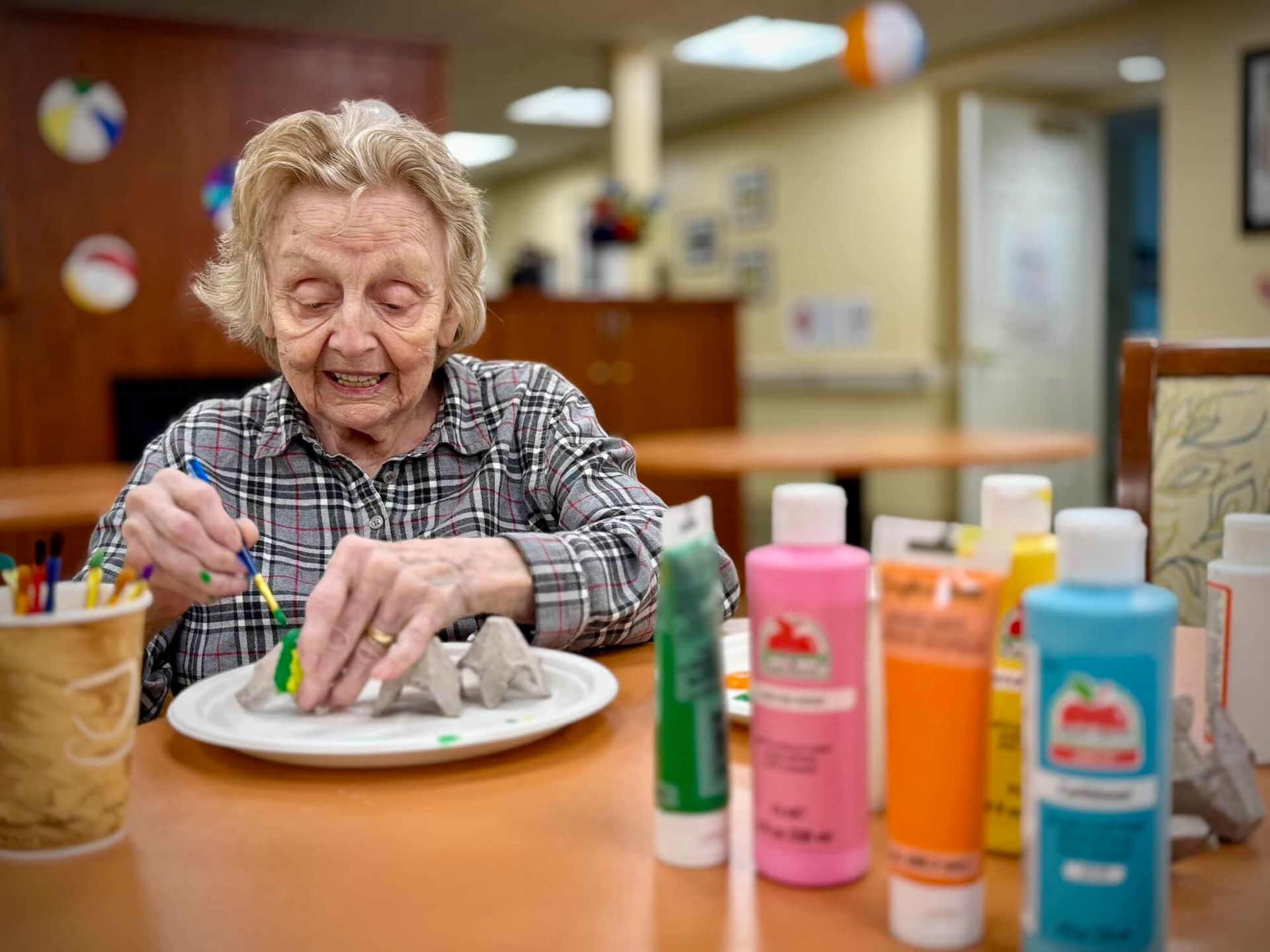 Woman painting green on a craft with paint tubes at a table. Beach balls decorate the background.