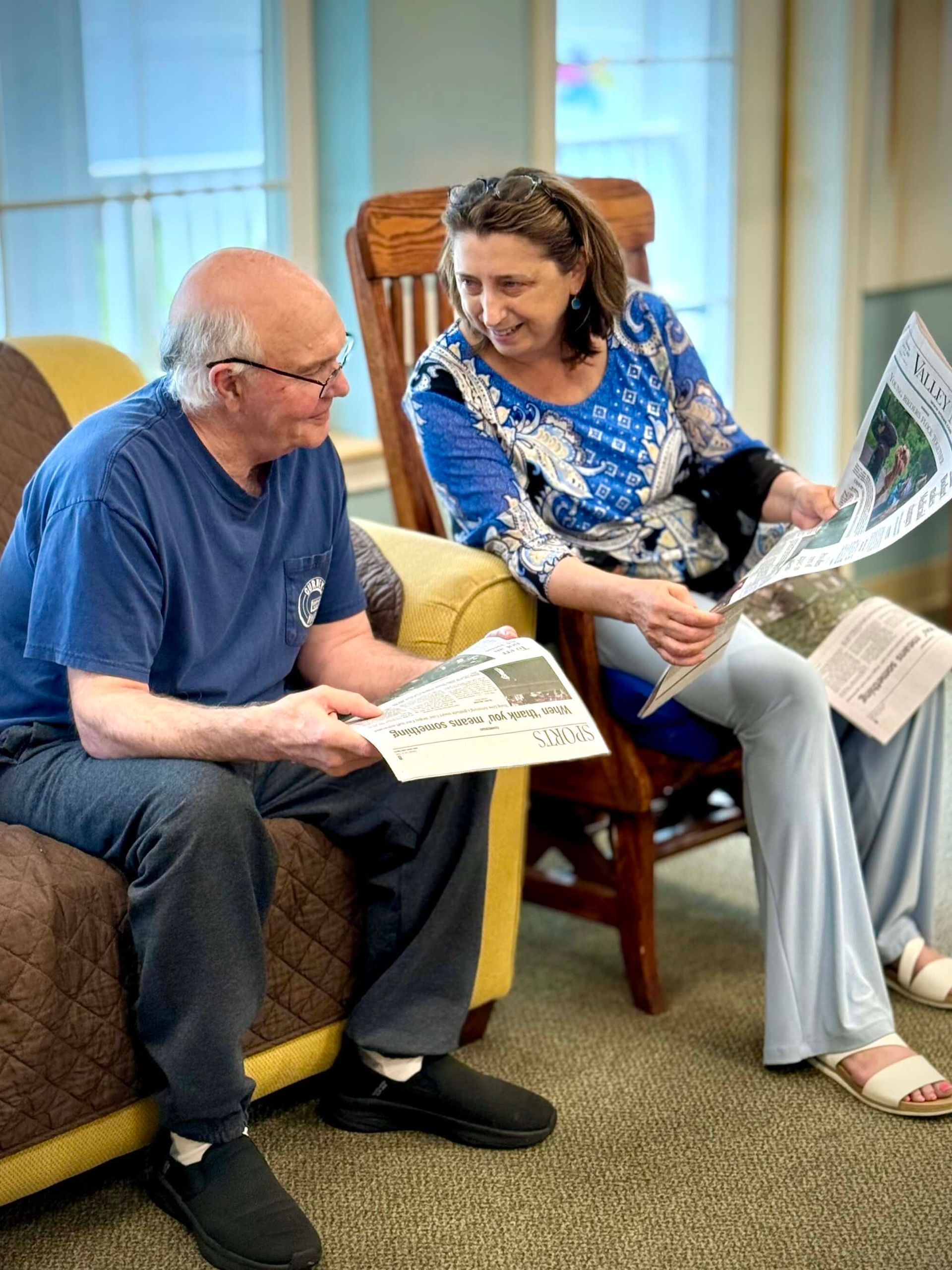 Two people seated, reading newspapers, smiling. Inside a room with a wooden chair and sofa.