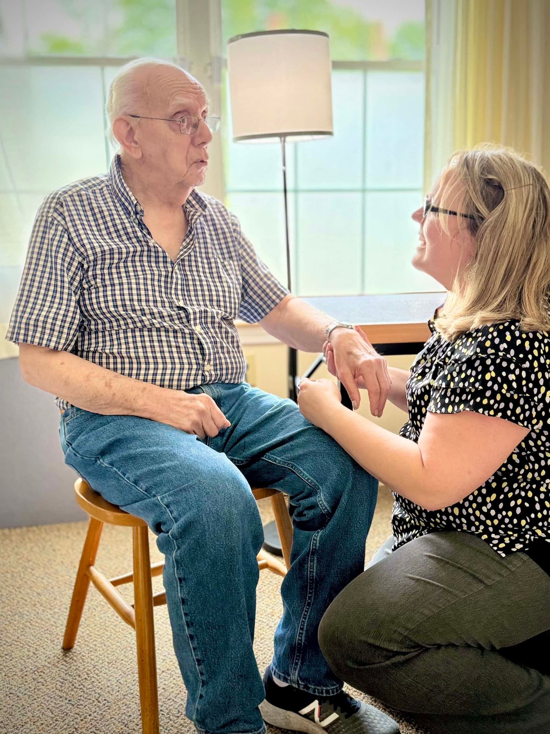 Man sits while woman checks his wrist indoors. Both are facing each other. Bright setting.