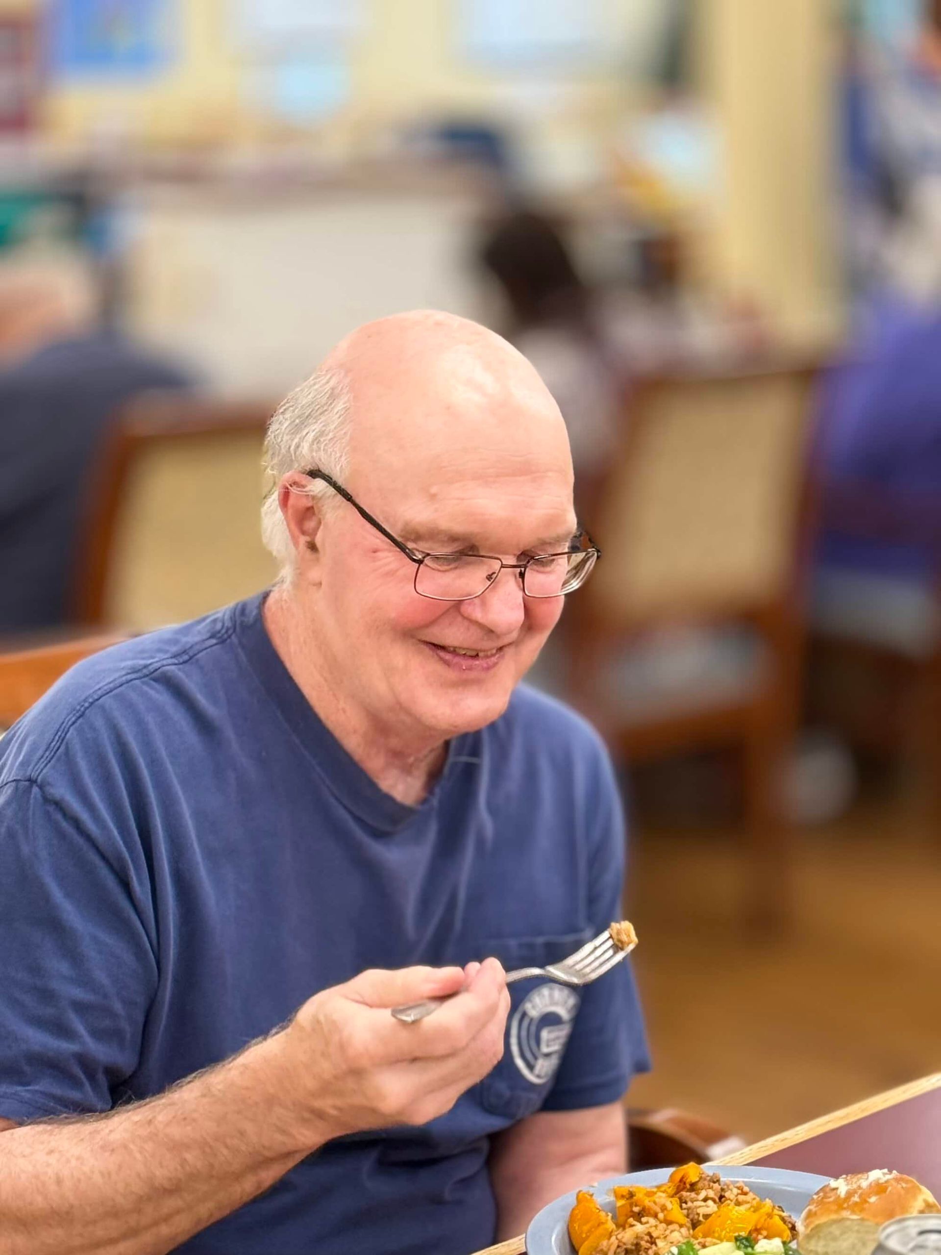 Bald man in glasses smiles while eating, indoors.