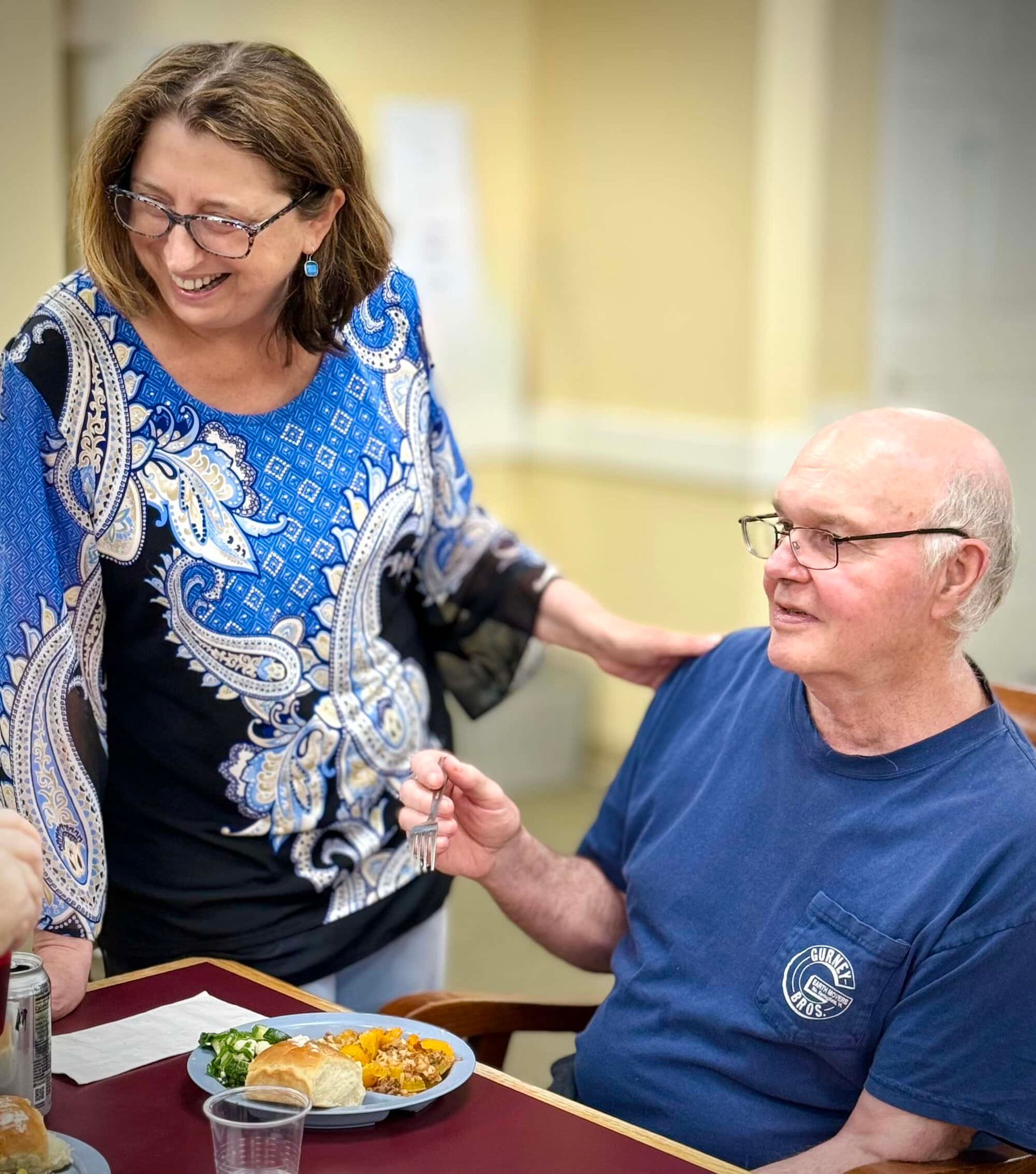 Woman in blue top touches the shoulder of a man in a blue shirt at a dining table; food and drink are present.