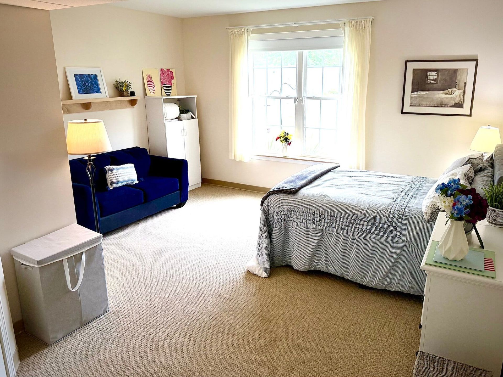 Bedroom with blue sofa, bed, white dresser, window, and neutral carpet.