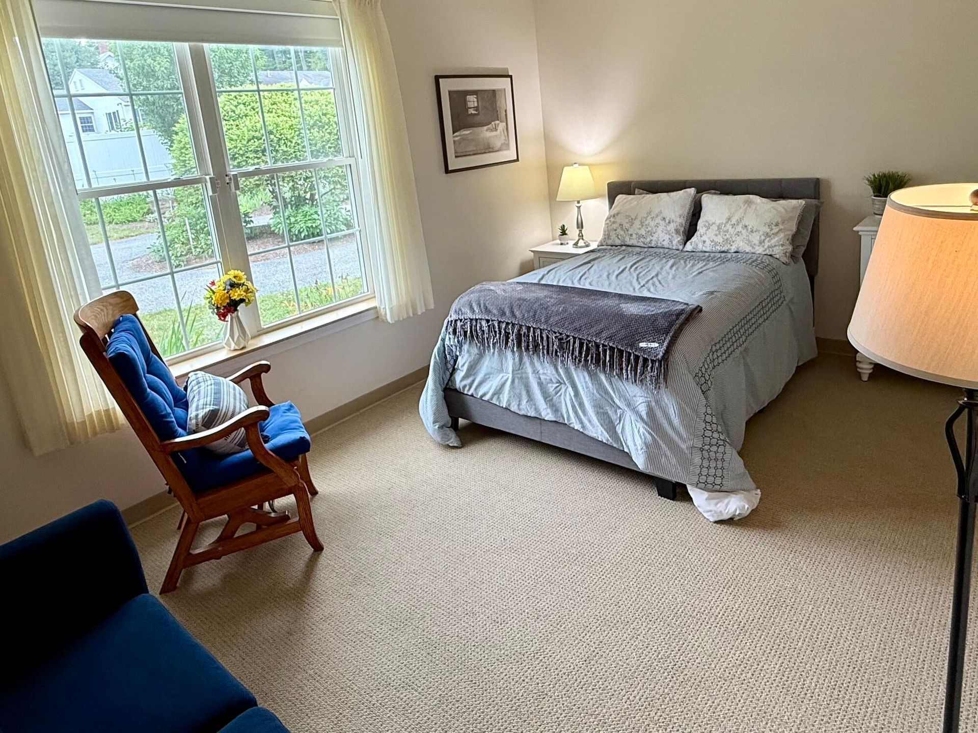 Bedroom with bed, rocking chair, window, lamp, and neutral carpet.