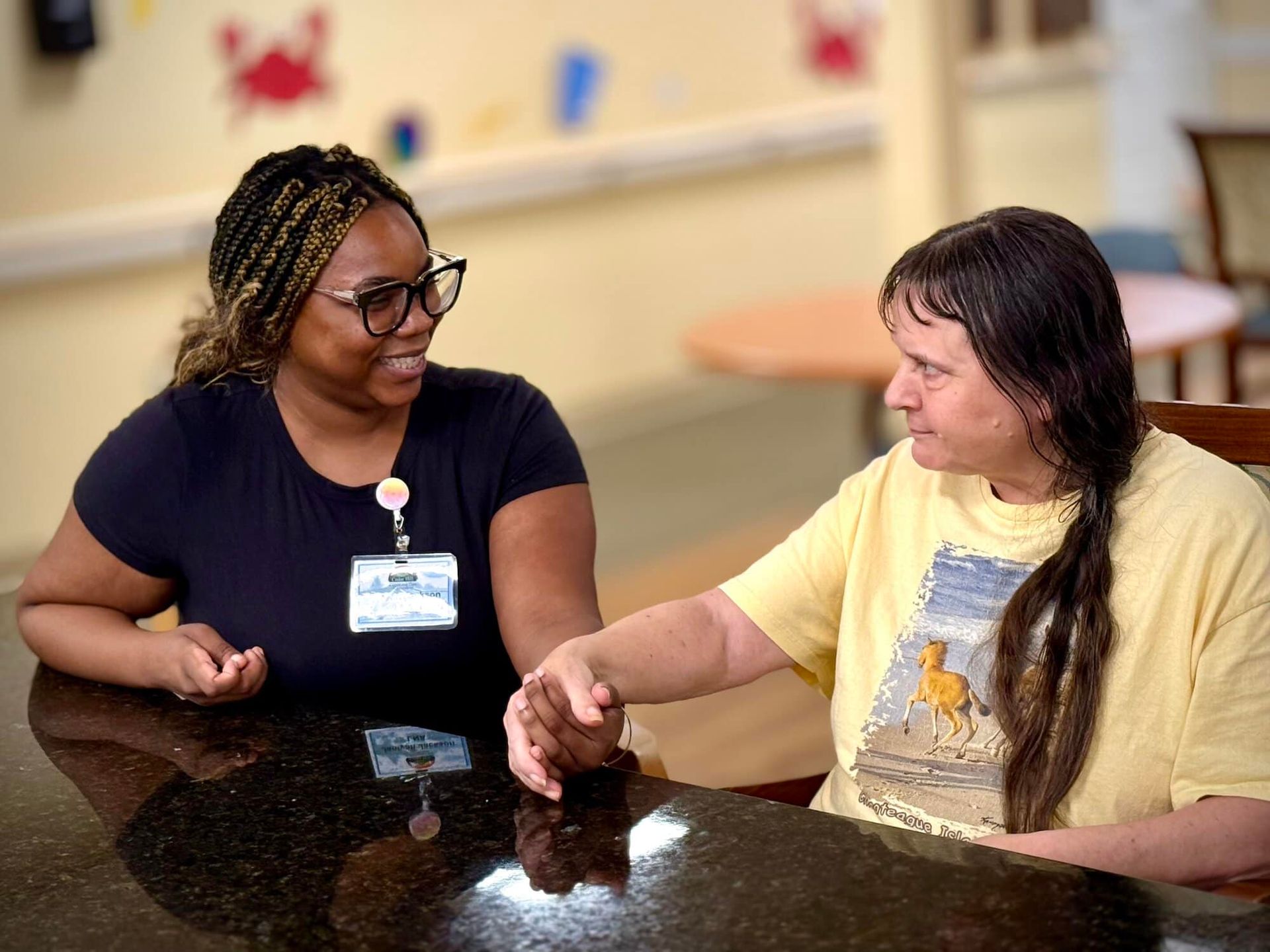 A person with braids and glasses smiles at another person holding their hand at a table.