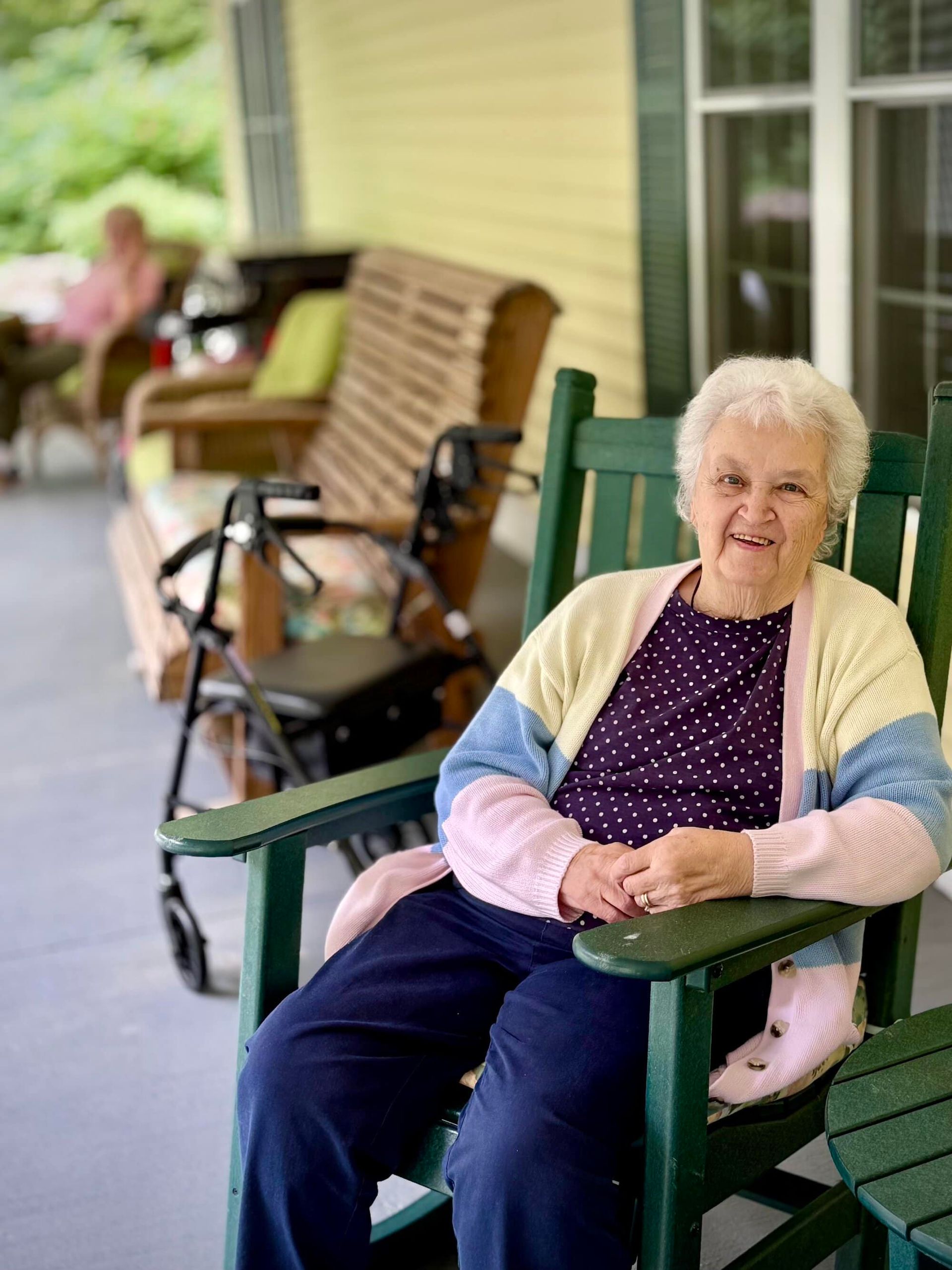 Elderly woman in green rocking chair on porch, smiling, light sweater, walker in background.