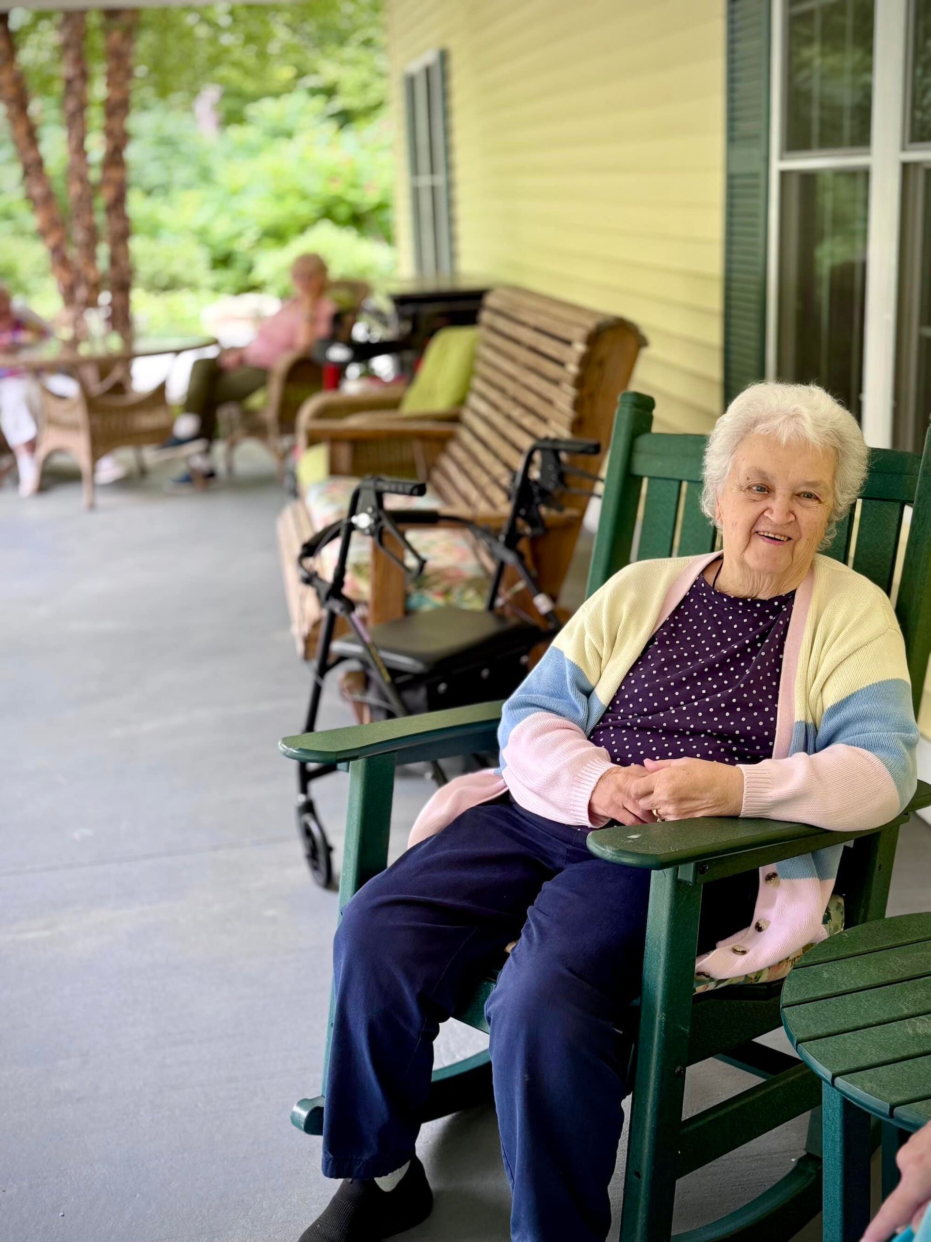 Elderly woman in a rocking chair on a porch, smiling. A second person is visible in the background.