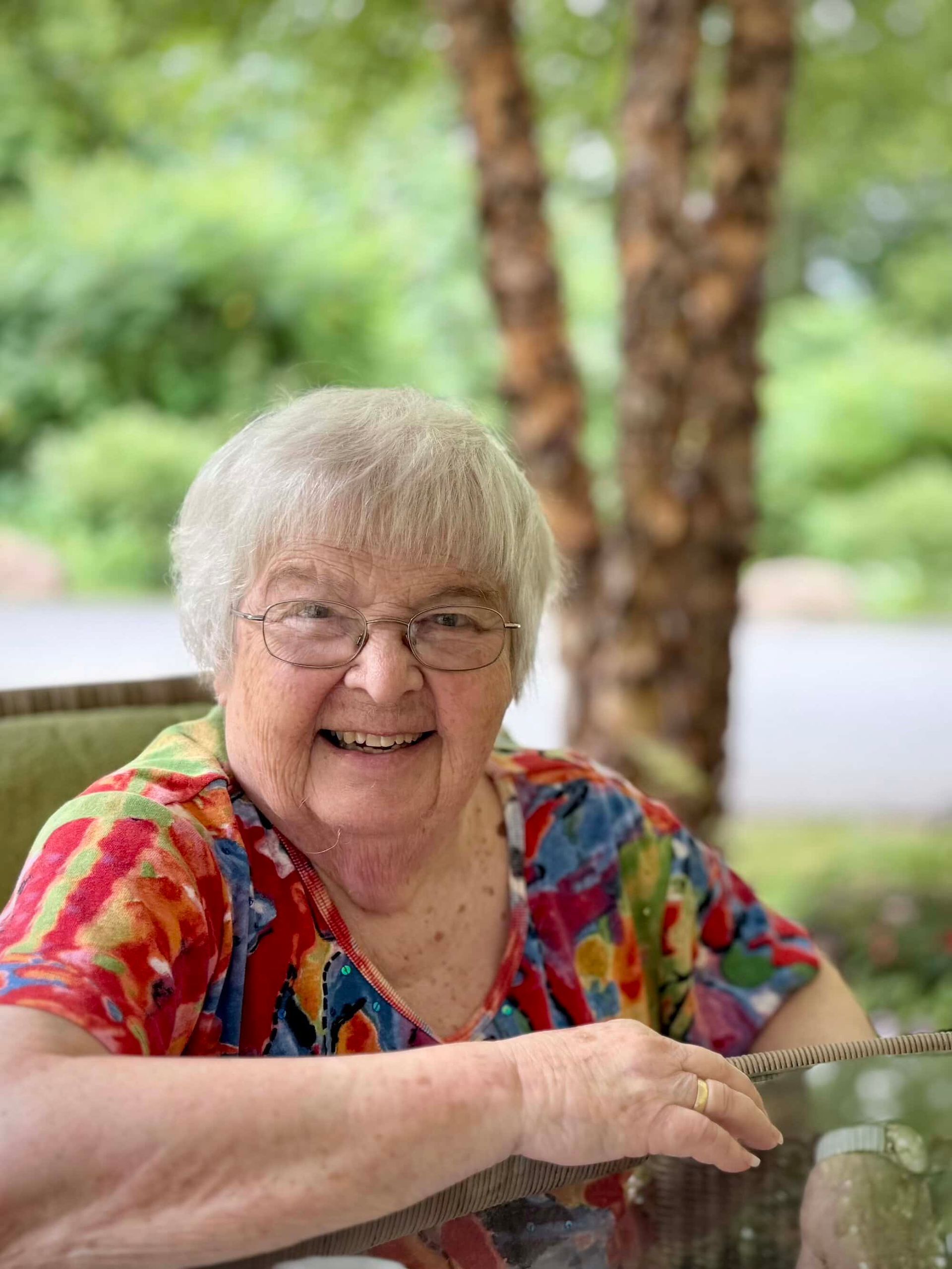 Smiling woman with glasses, colorful shirt, sitting outdoors.