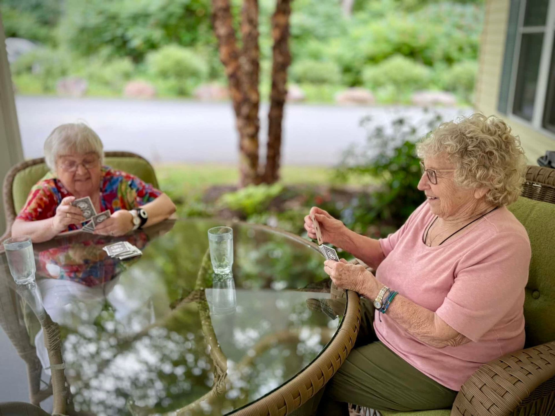 Two elderly women playing cards at a glass table on a porch.
