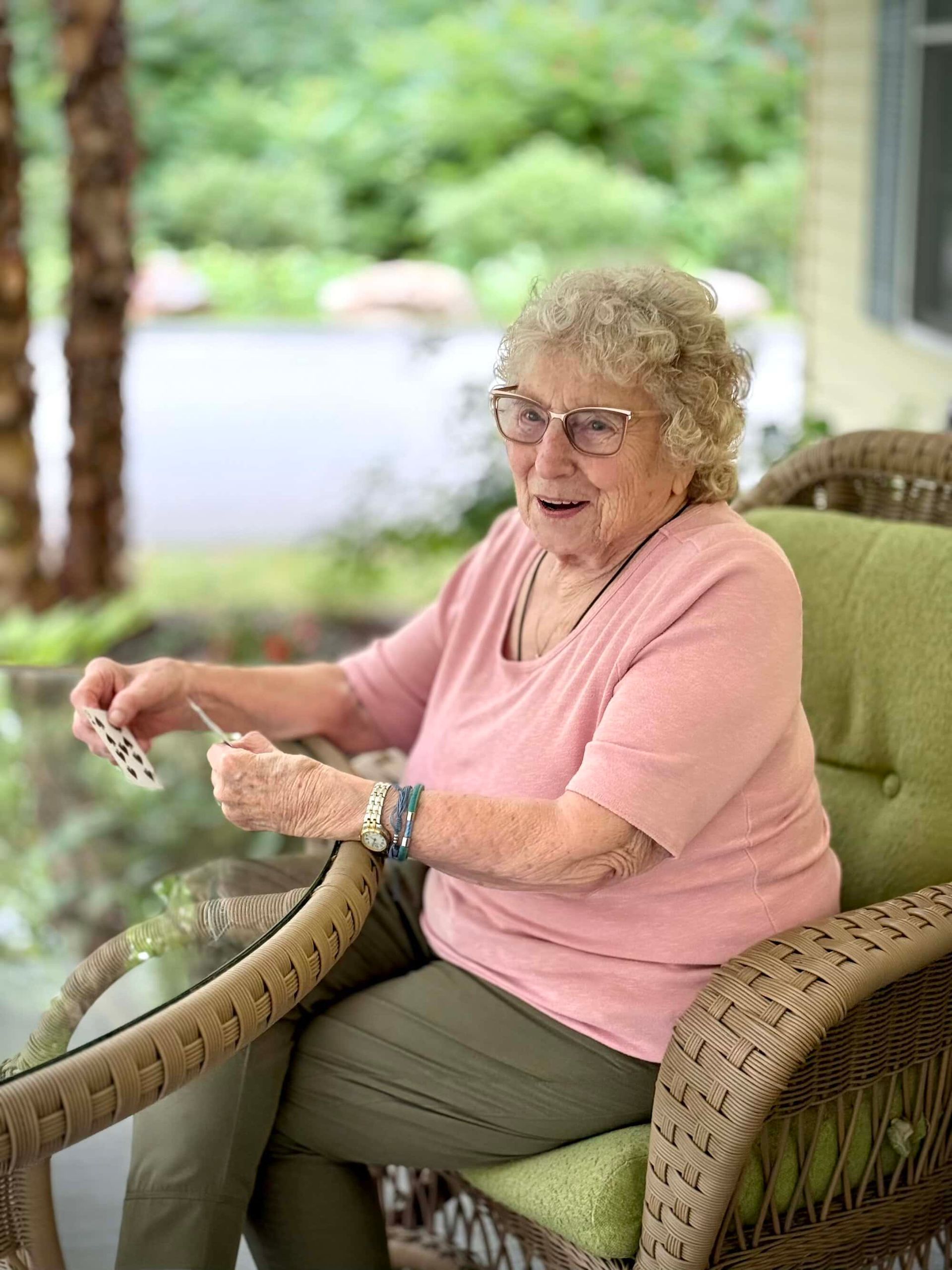 Elderly person with glasses sits in a wicker chair playing cards on a porch.
