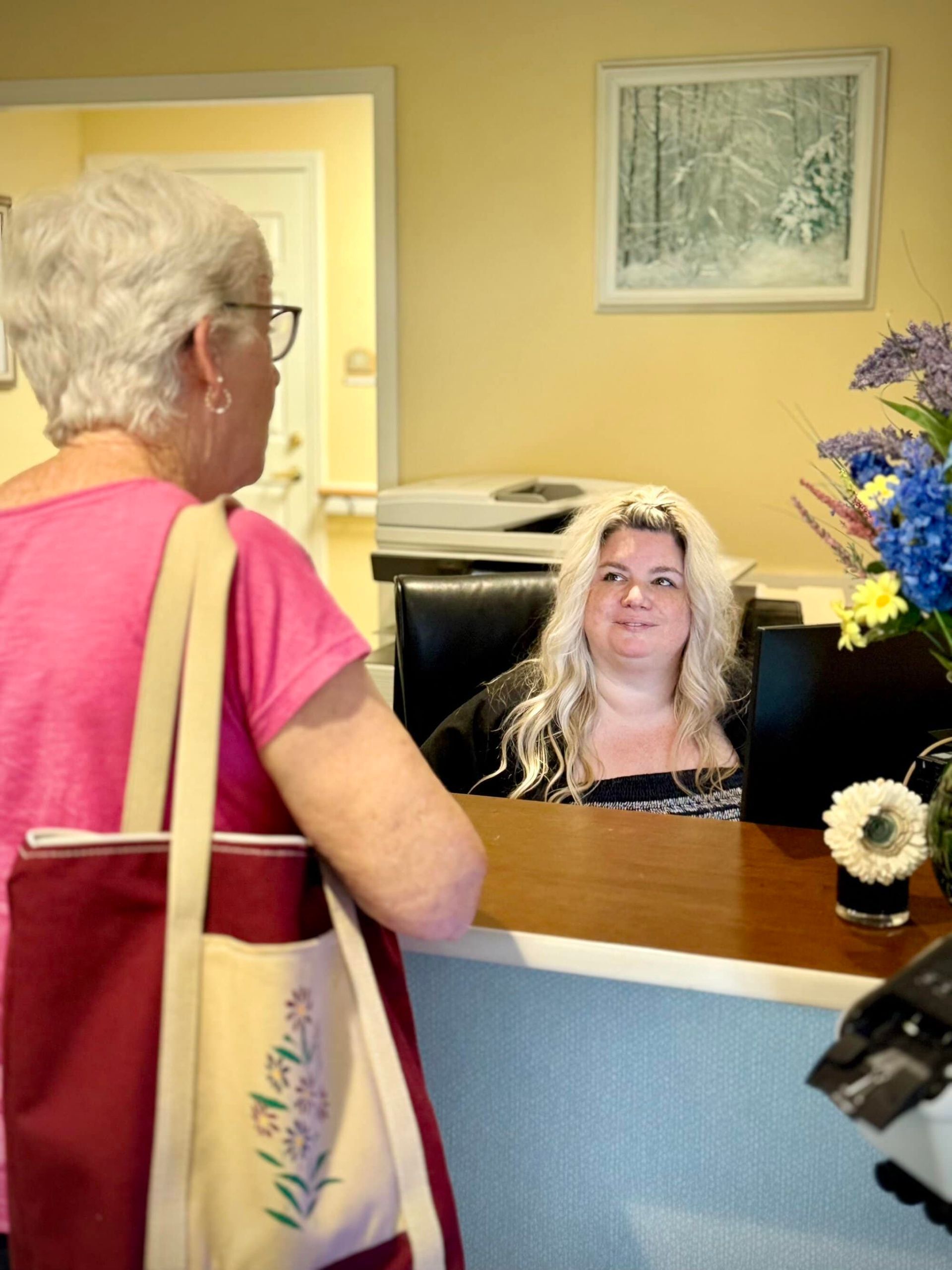 Woman with purse talking to receptionist at a desk. Flowers and a painting are visible.