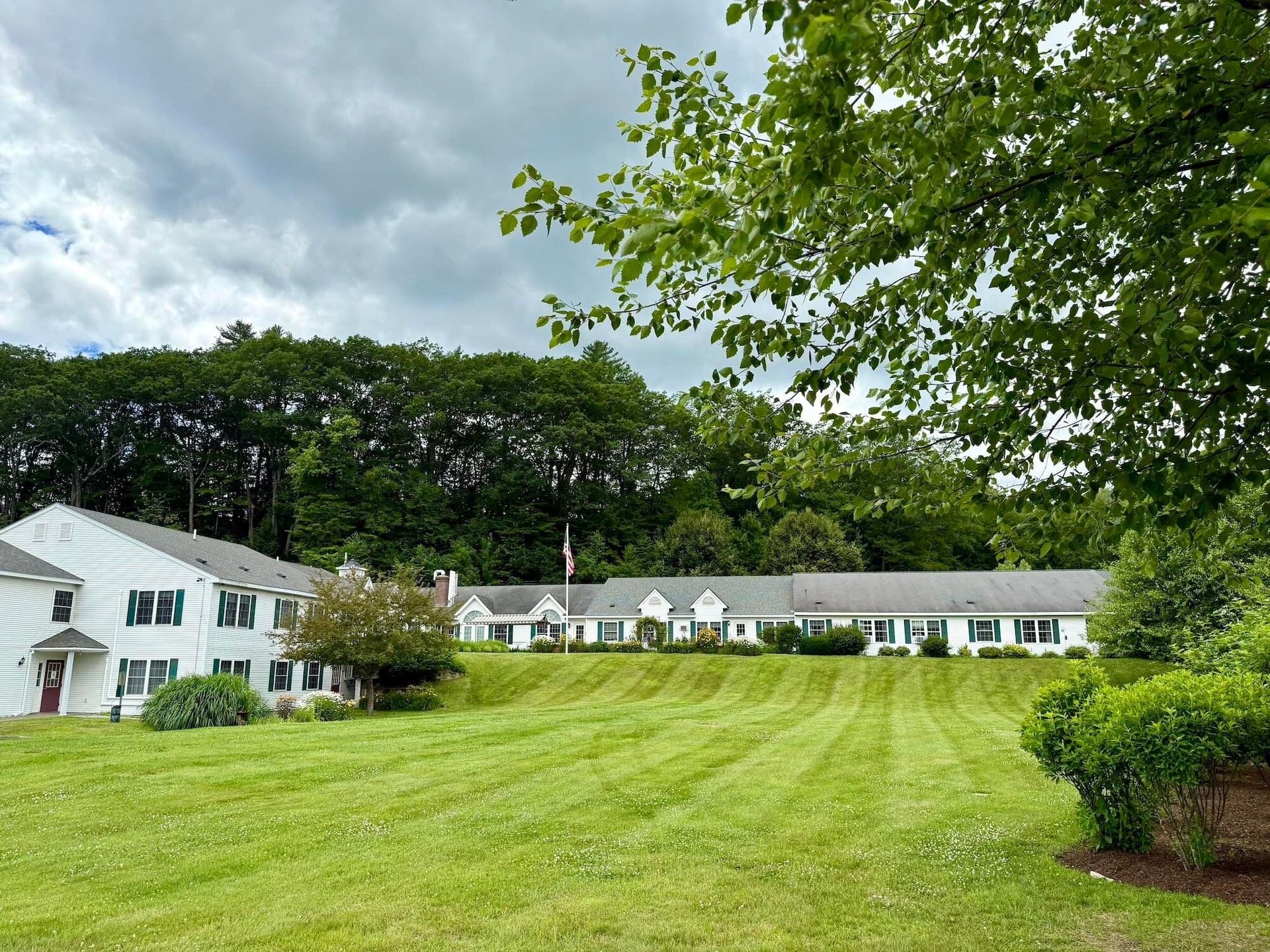 Lawn with long, low buildings and a taller white building. Trees in background, cloudy sky.
