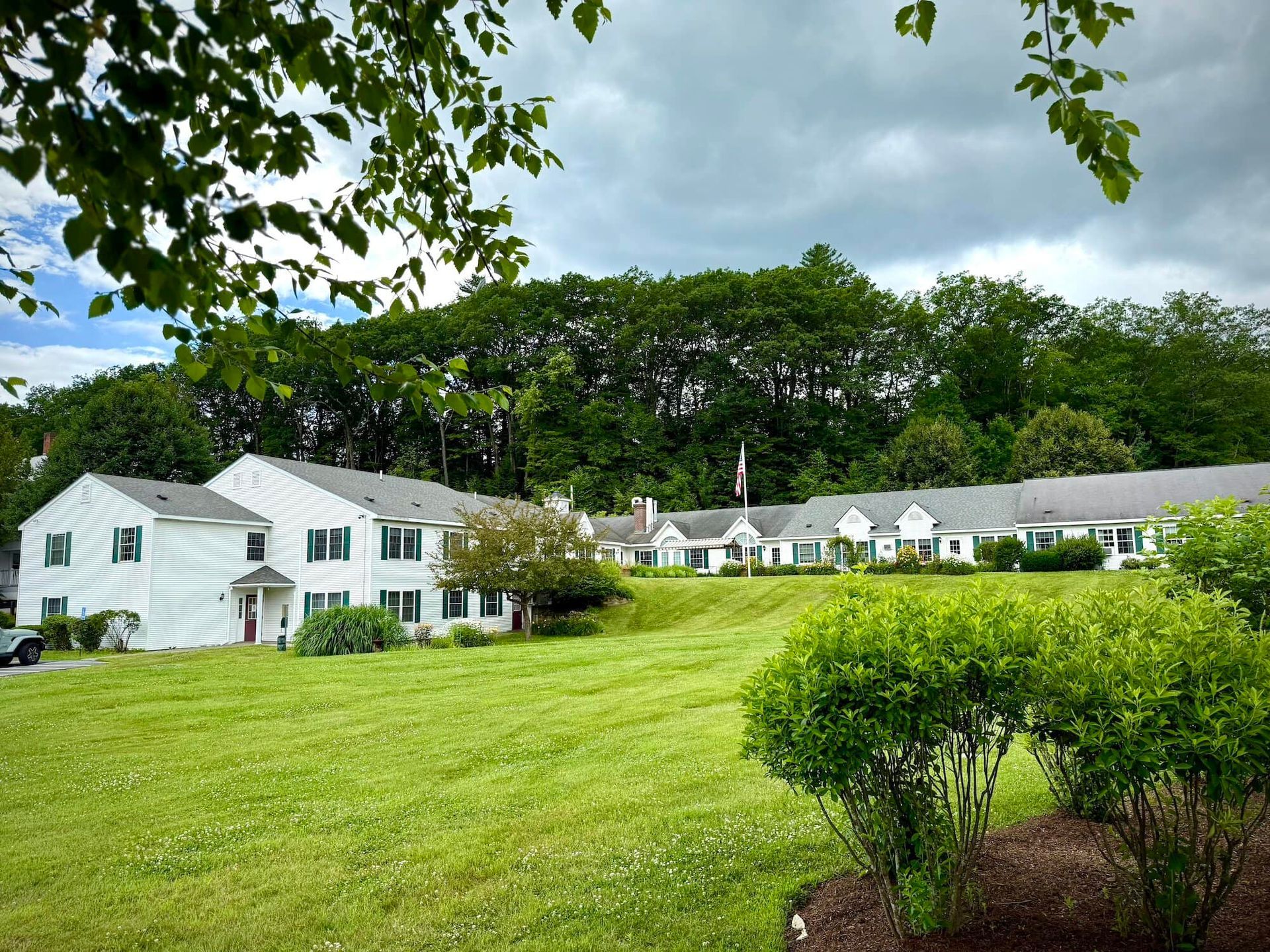 White buildings with green shutters sit on a grassy hill, backed by trees and a cloudy sky.