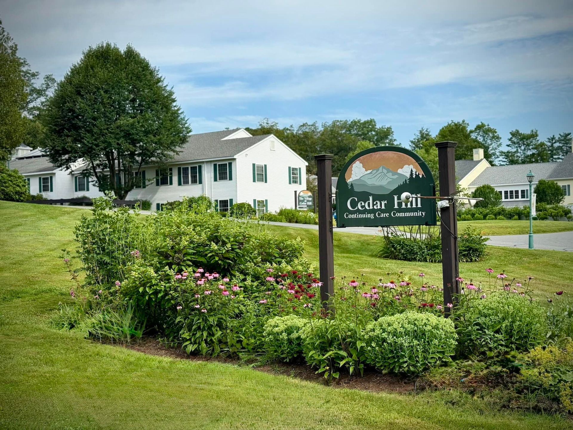 Cedar Hill sign in front of white buildings with green lawn and landscaping. Cloudy sky.