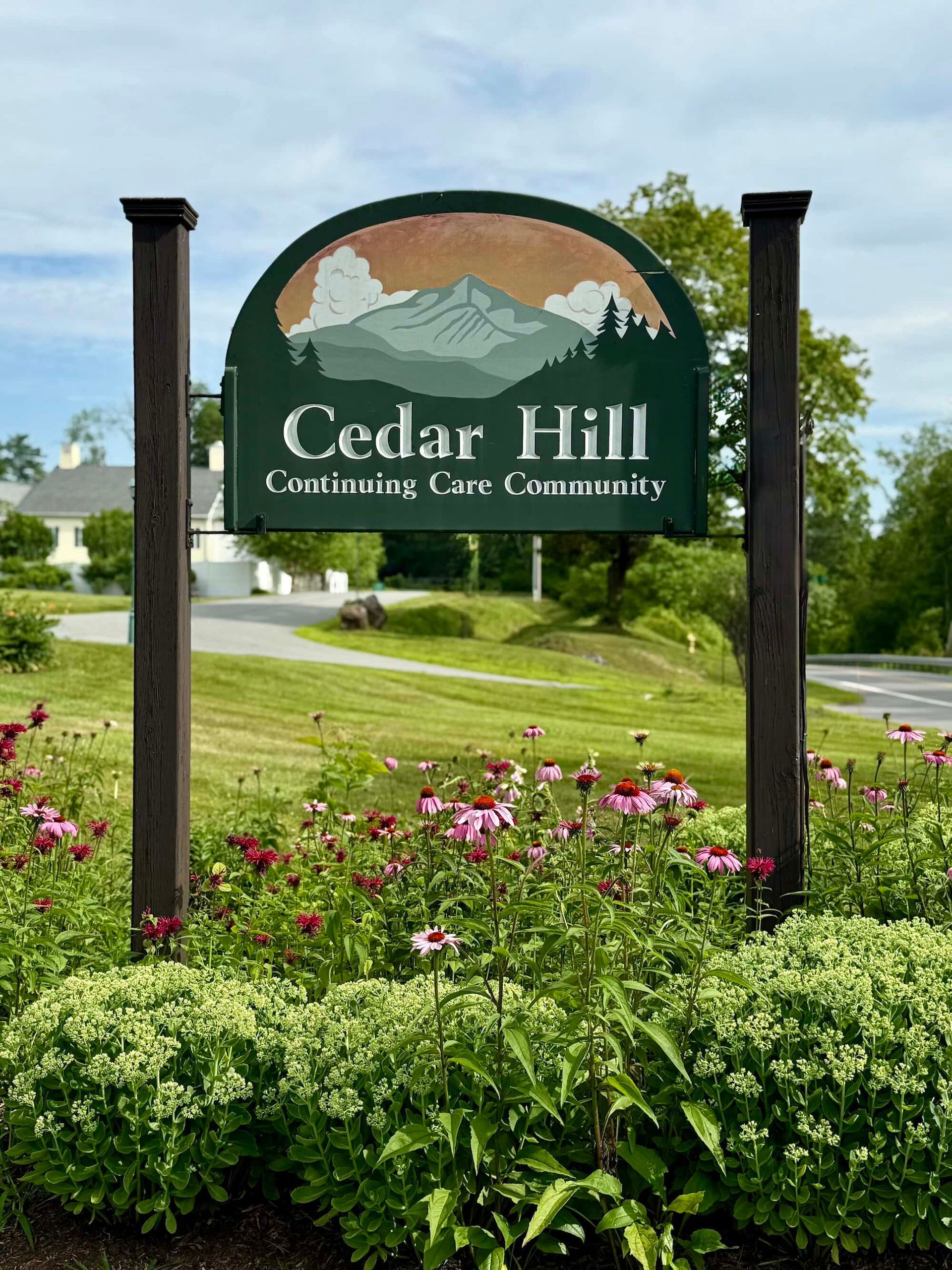 Sign for Cedar Hill Continuing Care Community, green with mountain scene, framed by brown posts, flowers in foreground.