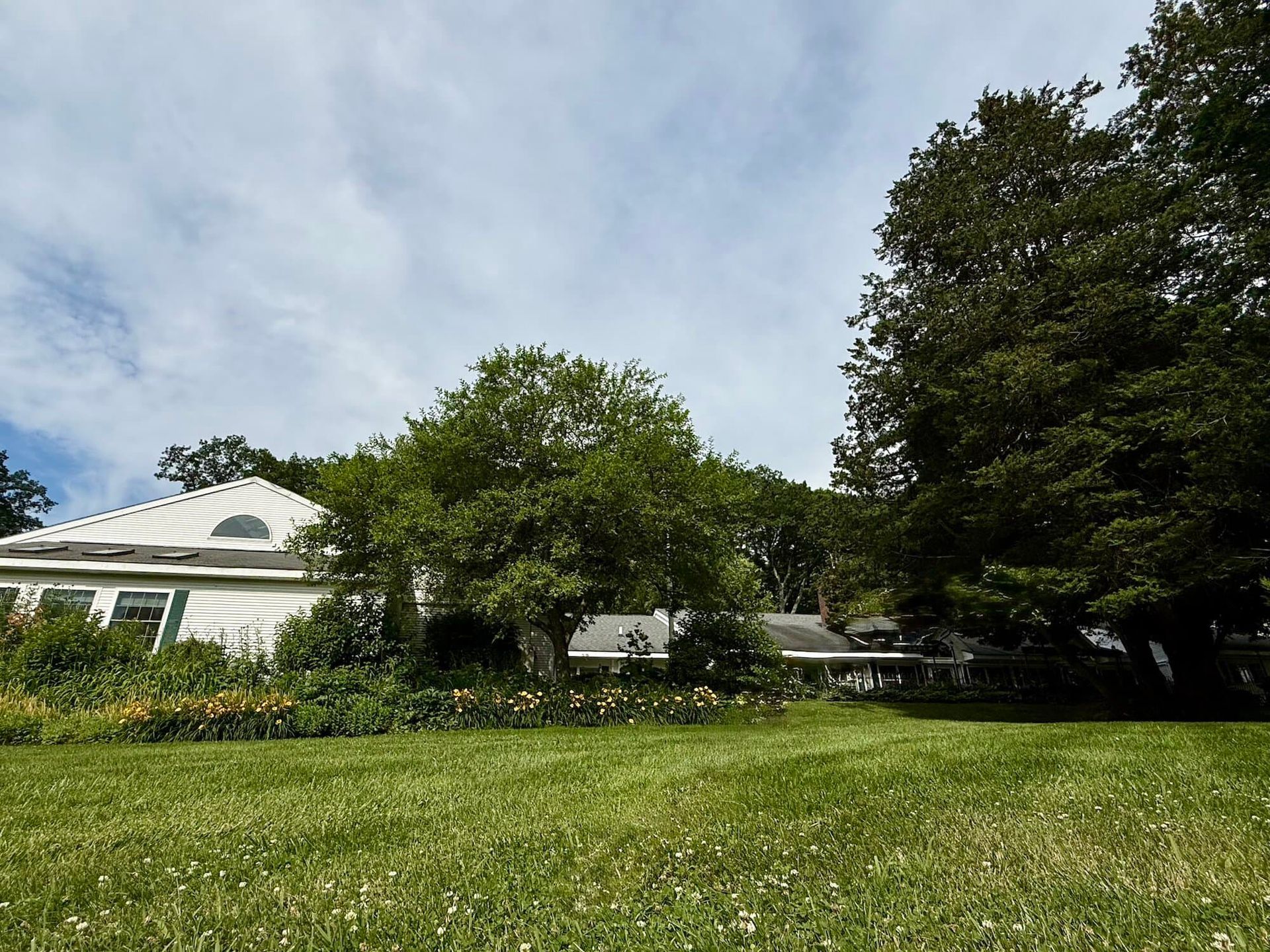 White house with a garden and large trees under a partly cloudy sky.